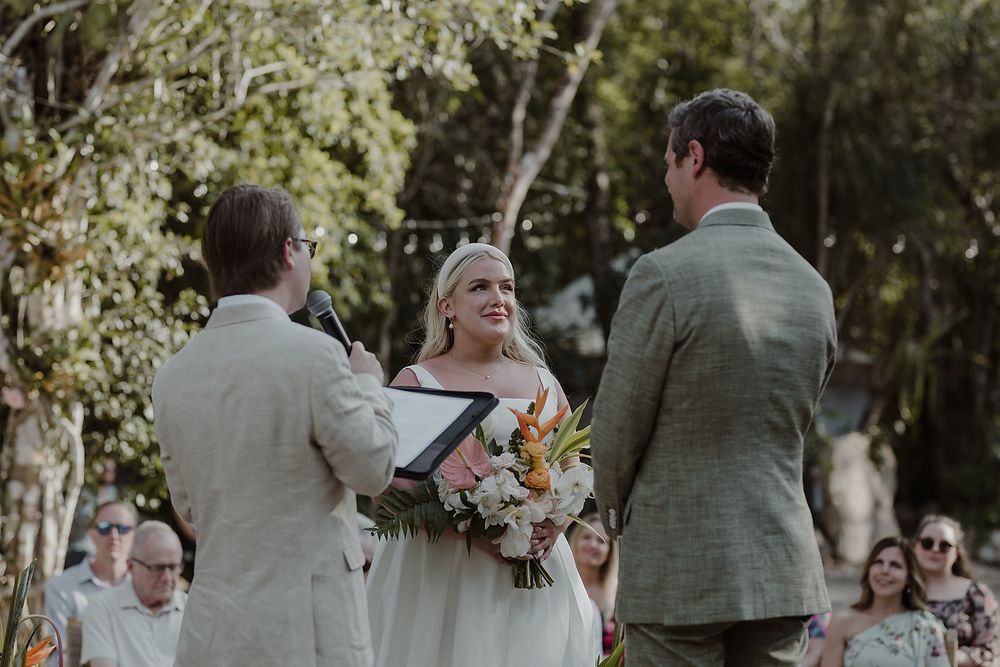 Bride holding floral bouquet smiles at groom during outdoor wedding ceremony with officiant reading vows.