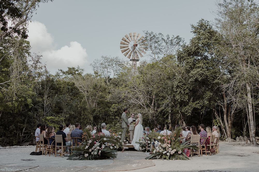 Outdoor wedding ceremony in a lush forest clearing with guests seated, floral arrangements, and a rustic windmill backdrop.