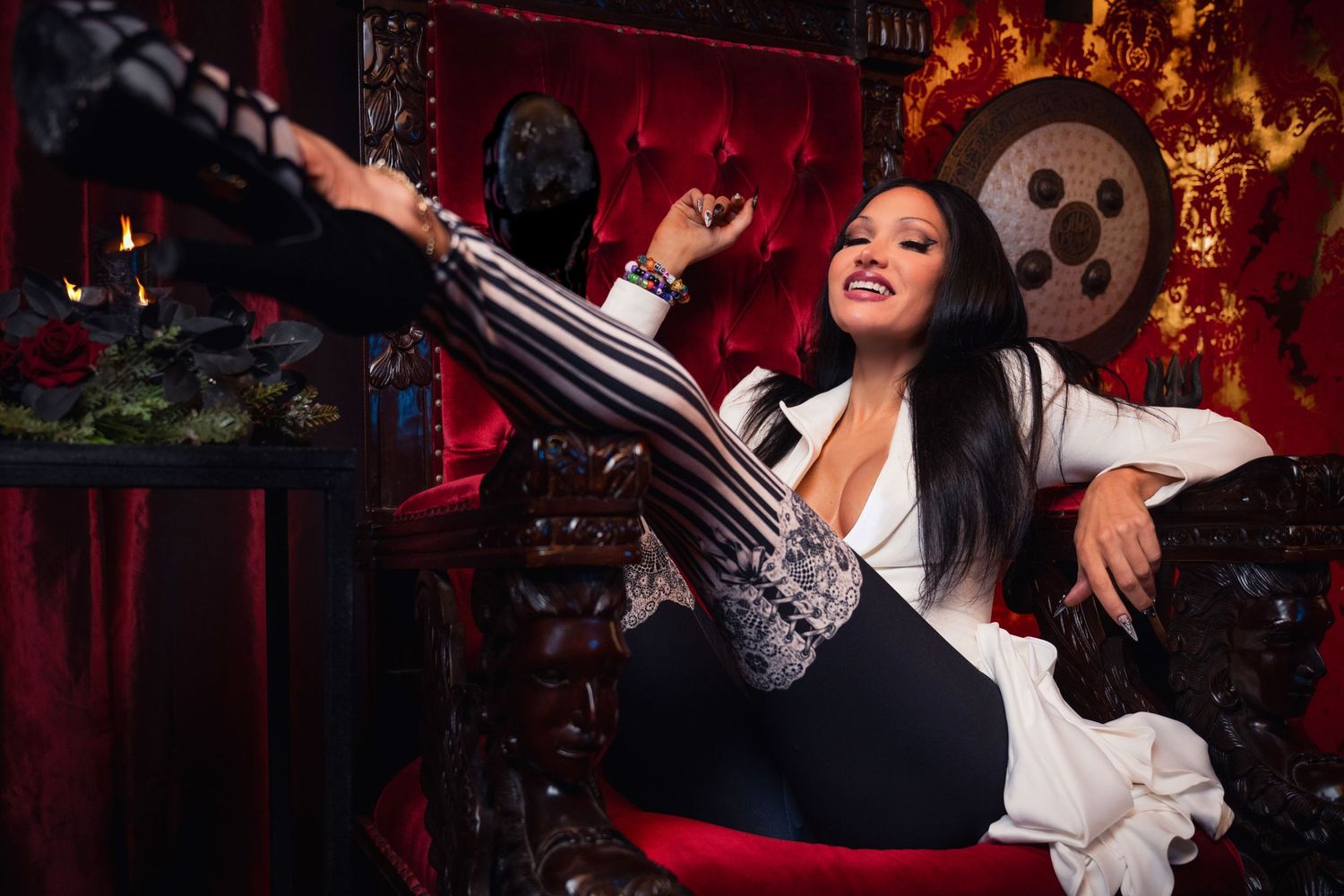 A figure lounges on a gothic throne with a chainsaw prop surrounded by spooky Halloween decorations and red lighting.