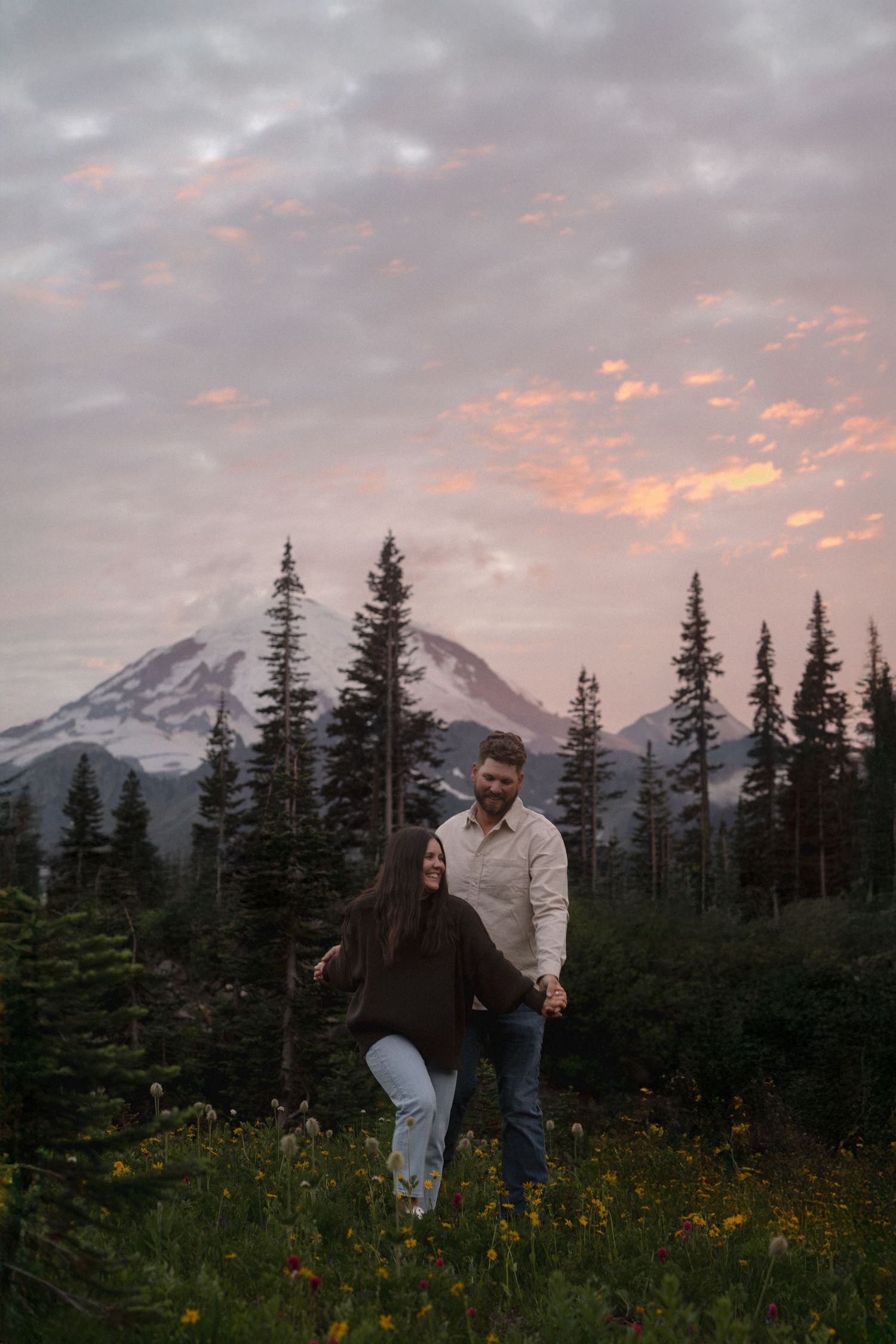 Couple stands in wildflower meadow at sunset with Mount Rainier and evergreen trees silhouetted in the background.