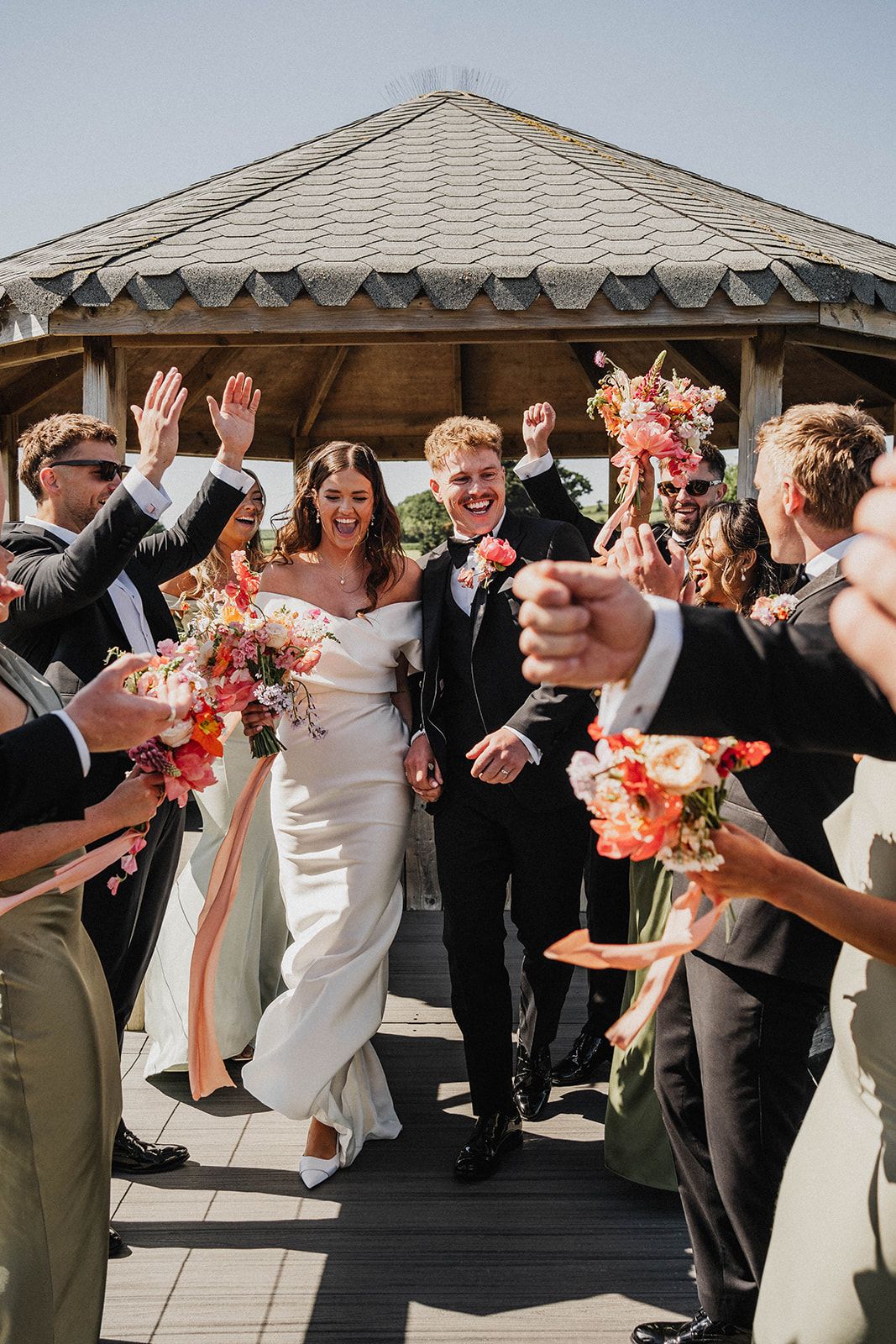 Joyful wedding celebration with bridal party in formal attire cheering under outdoor gazebo with coral floral bouquets.