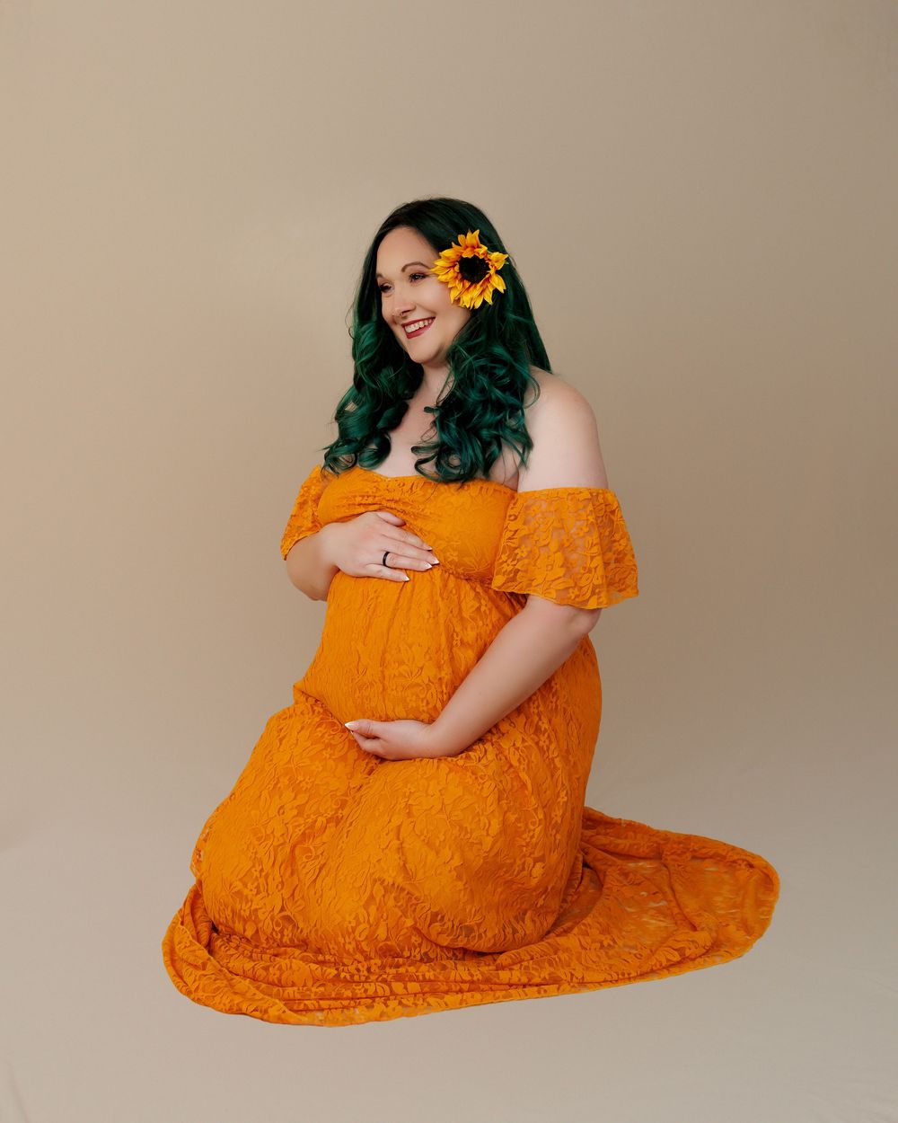 A pregnant woman wearing an orange lace off-shoulder dress sits elegantly with a sunflower in her green hair.