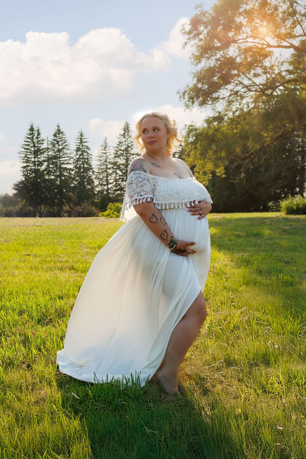 Woman in flowing white dress twirls gracefully in sunlit meadow with evergreen trees in background.