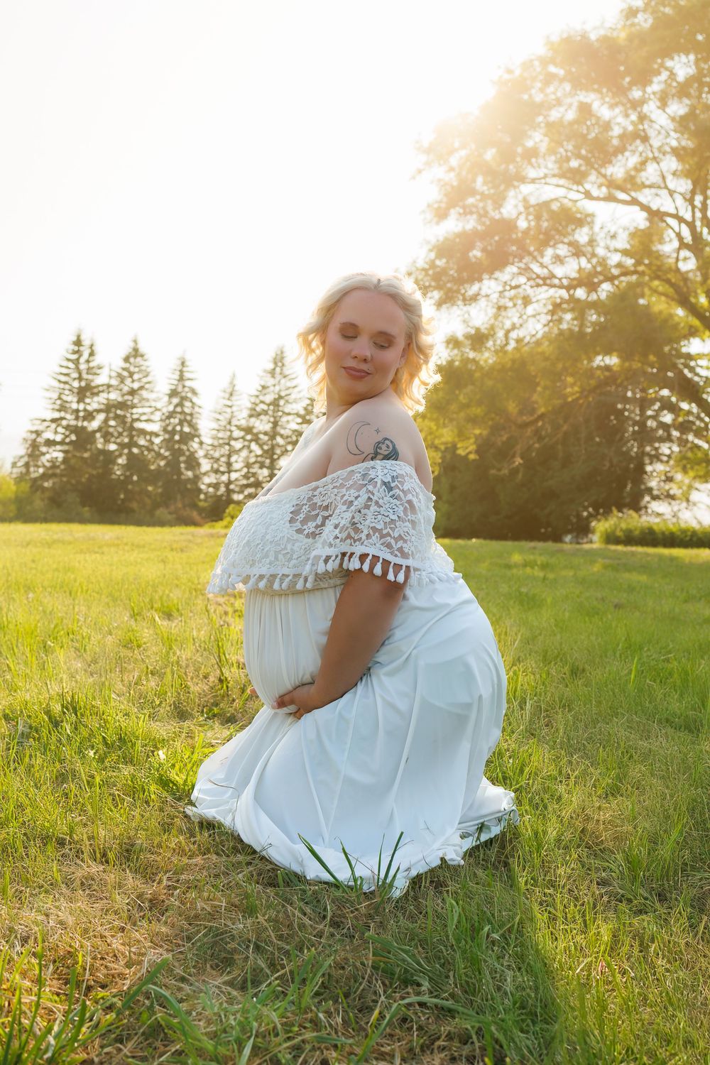 Ethereal maternity photo in a lace white dress at sunset in a grassy meadow with evergreen trees in background.
