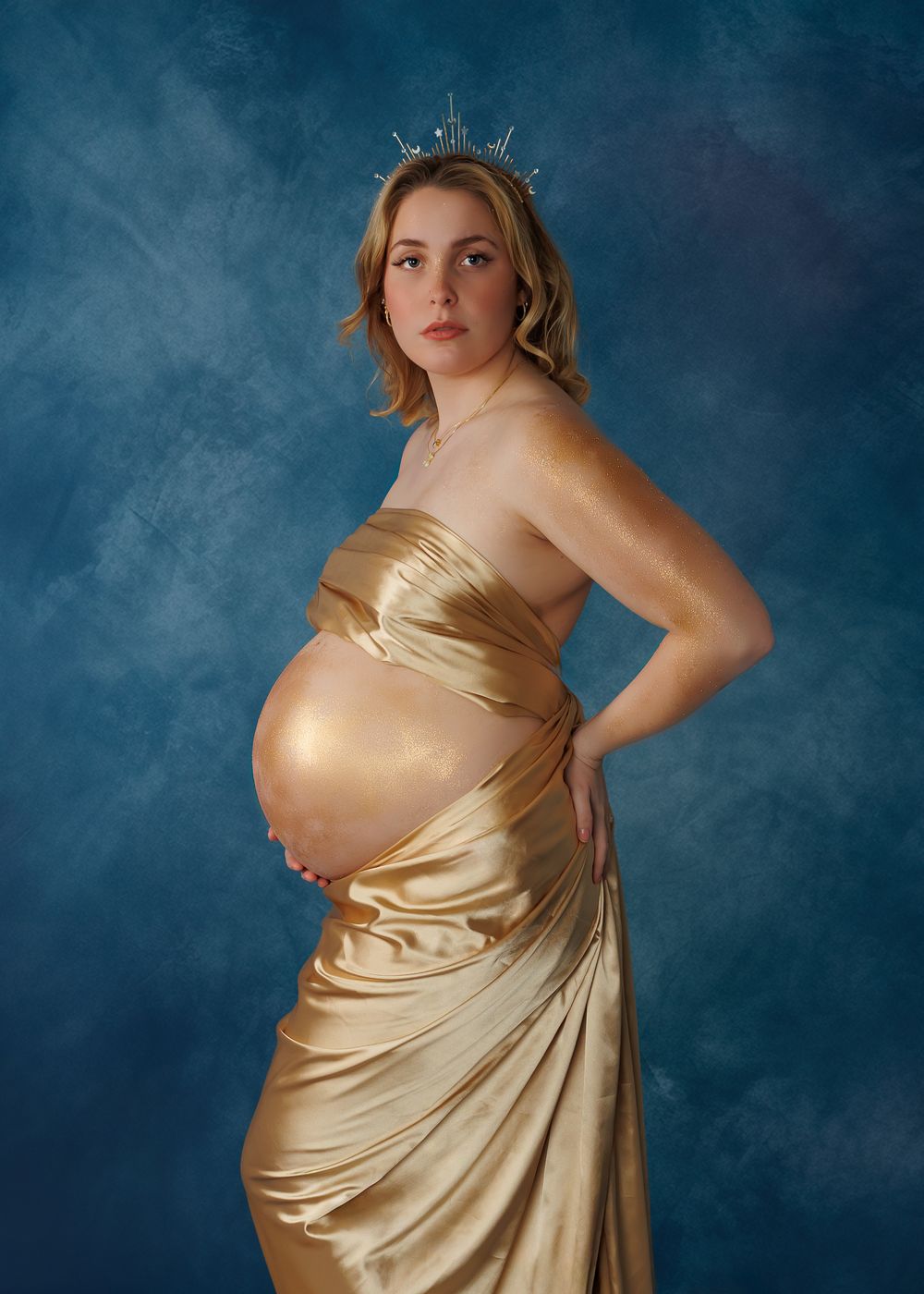 Someone wearing an elegant gold satin dress poses against a moody blue backdrop in a series of maternity photos.
