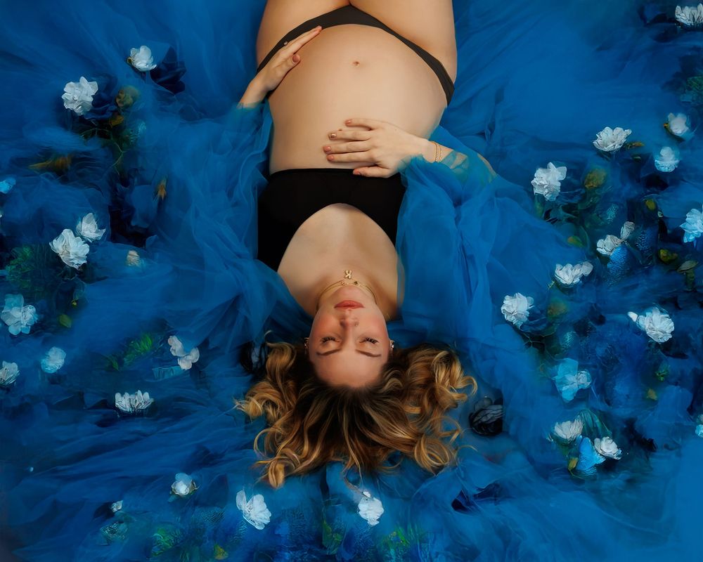 Person with flowing hair lying on blue fabric surrounded by scattered white flowers in an ethereal portrait setup.