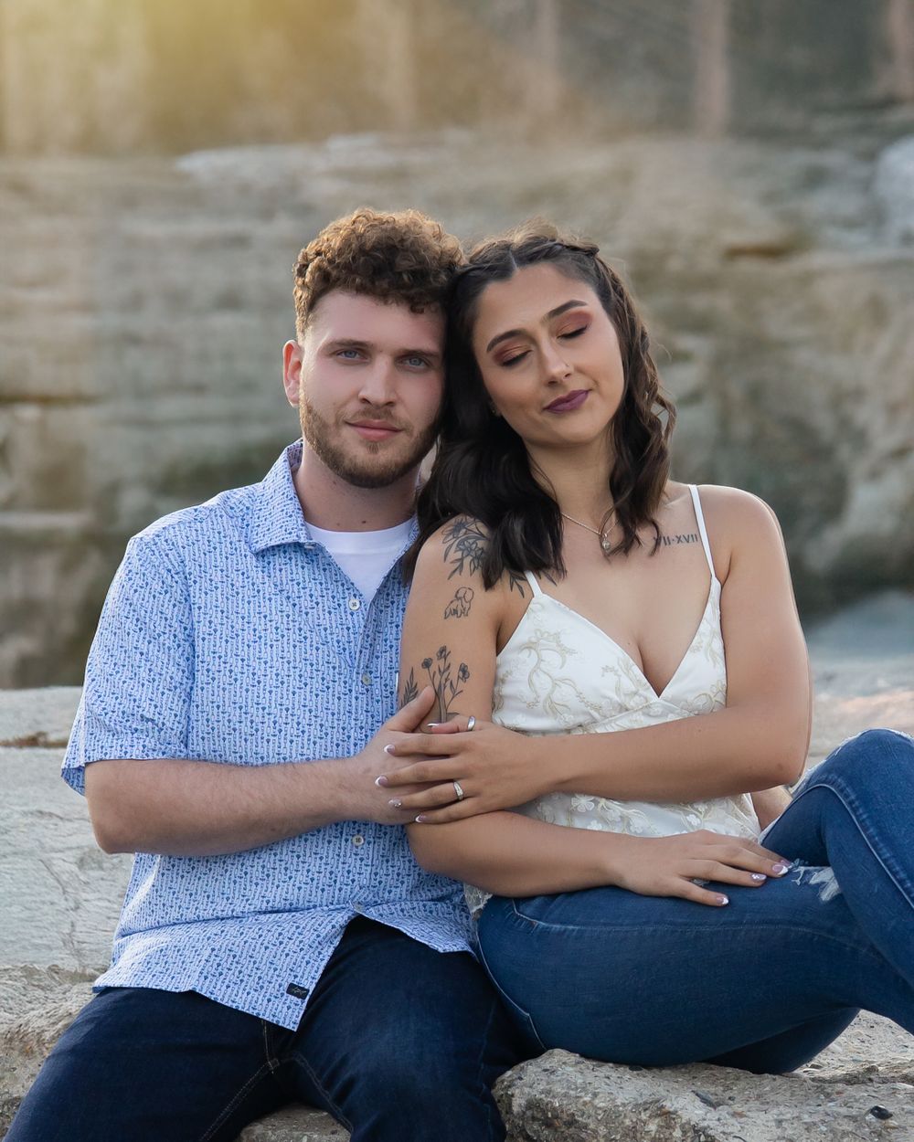 A couple sits intimately together on rocky terrain during a romantic outdoor photo session at golden hour.