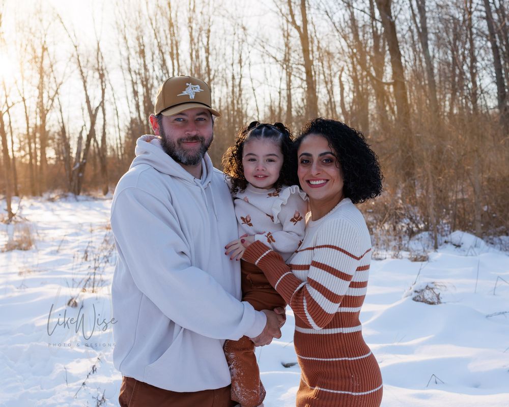 A family poses together on a snowy winter day in the woods at sunset.