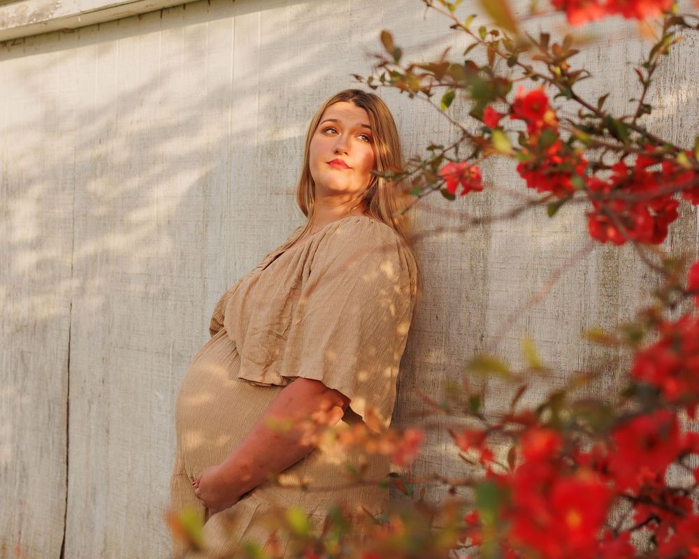 Woman in earth-toned sweater poses outdoors near vibrant red autumn flowers against a light colored wall.