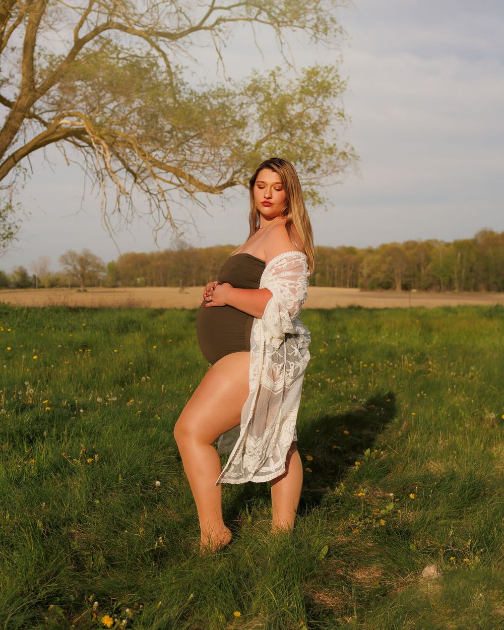 A maternity photo shows a figure in a white lace robe standing in a golden field with wildflowers at sunset.