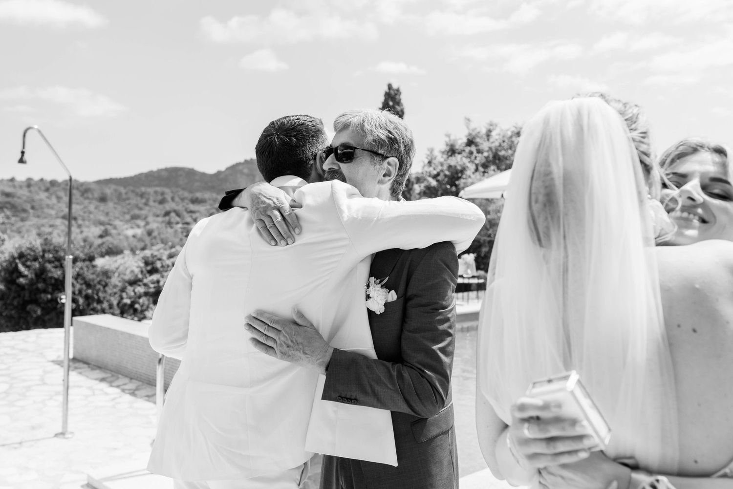 A black and white photo of people embracing at an outdoor wedding celebration with mountains in the background.