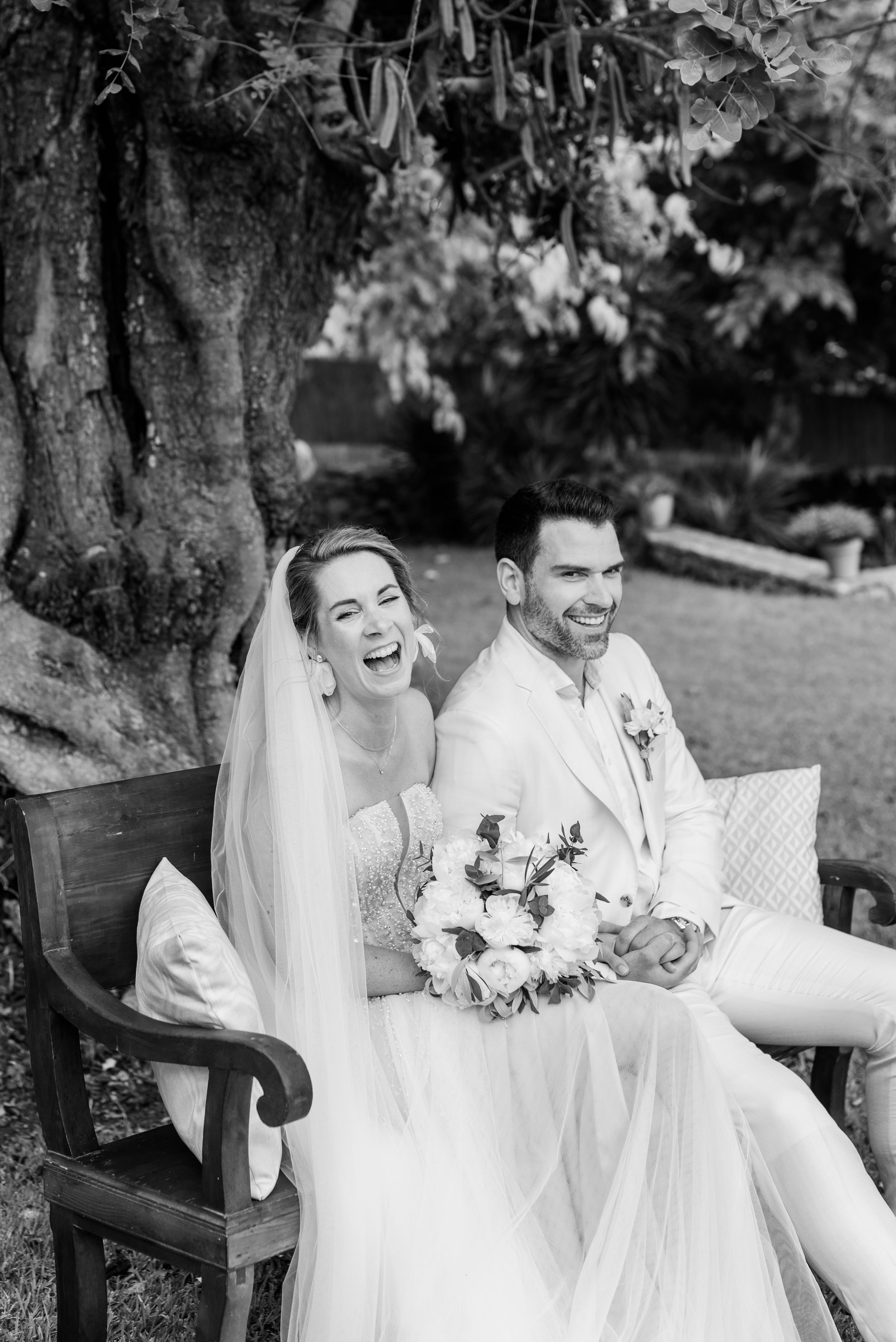 Ein Paar in weißer Hochzeitskleidung sitzt auf einer Bank und teilt auf der Wiese der Finca Xiclati herzliches Lachen und freudige Momente.