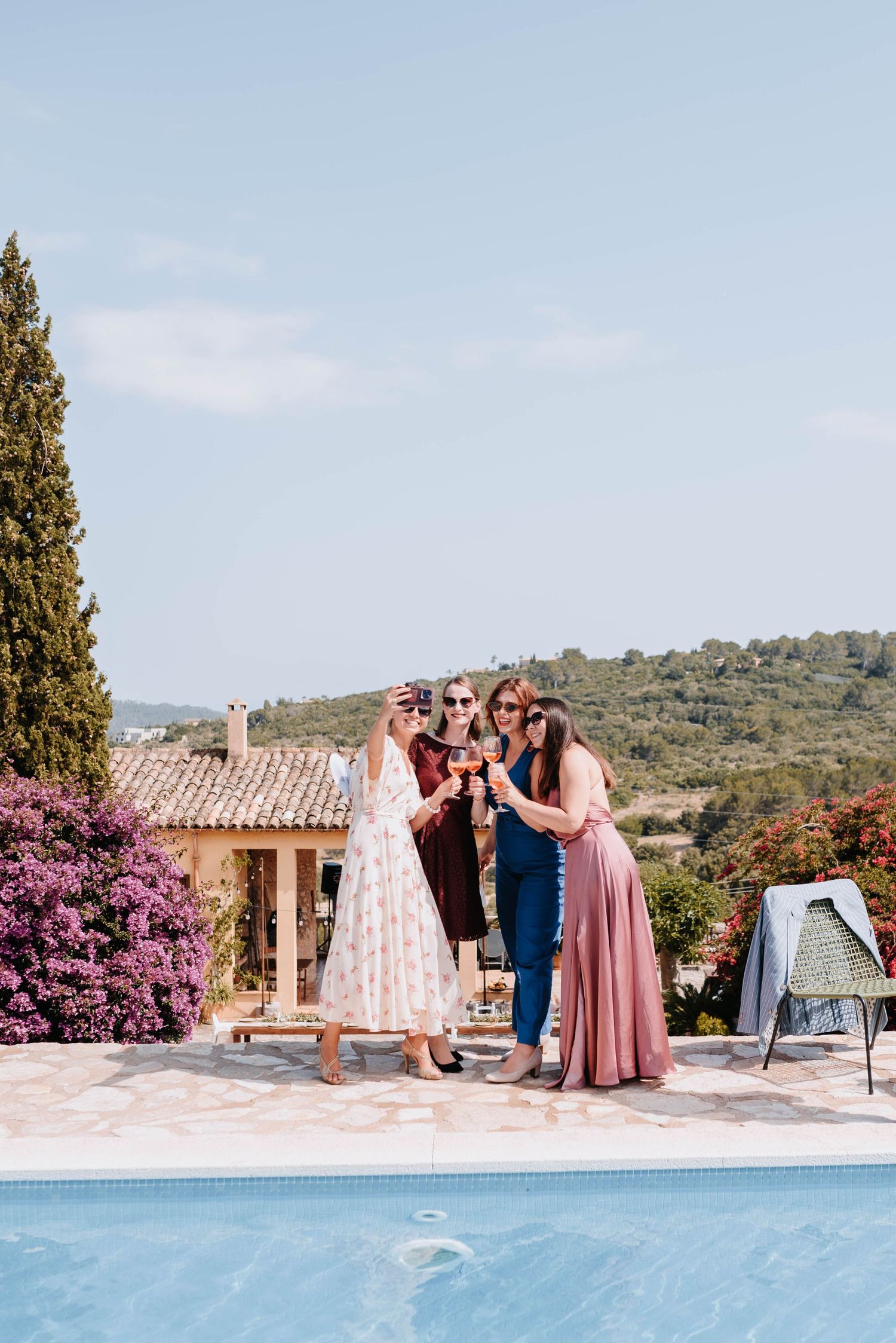 Gäste einer Hochzeit in formeller Kleidung versammeln sich an einem Swimmingpool in einer mediterranen Landschaft in hellen Pastellfarben.