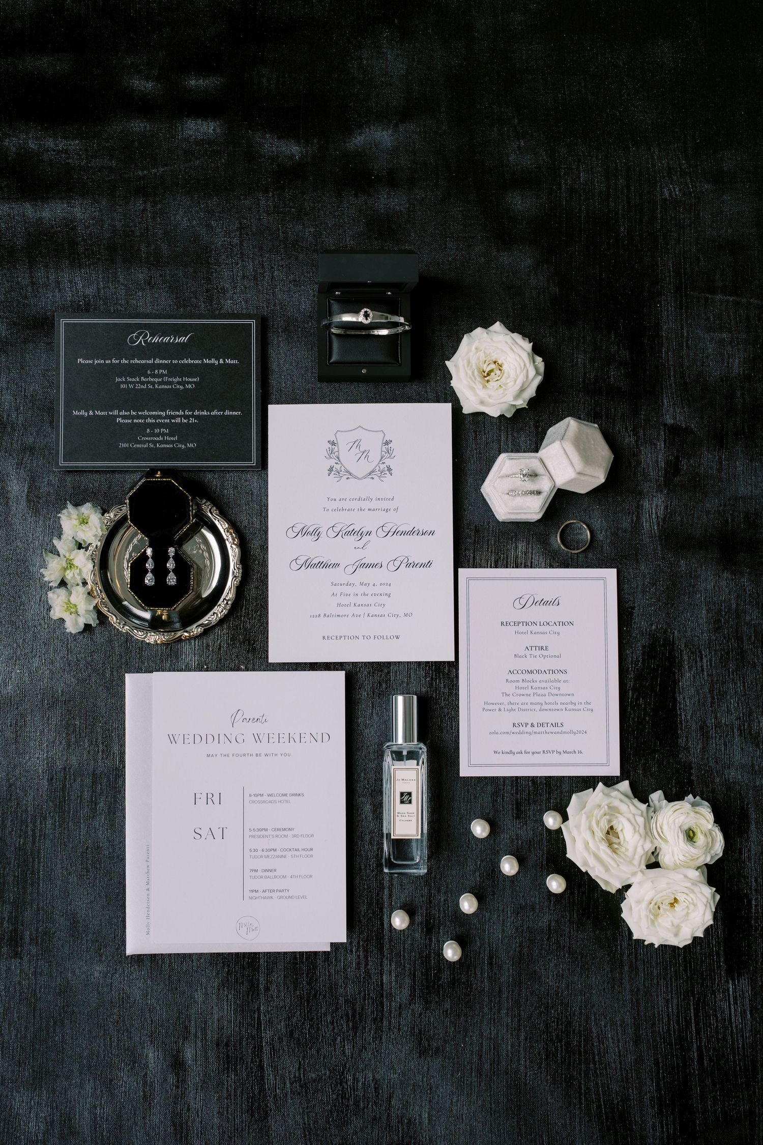 Monochrome wedding invitation suite with white flowers and perfume on dark surface.