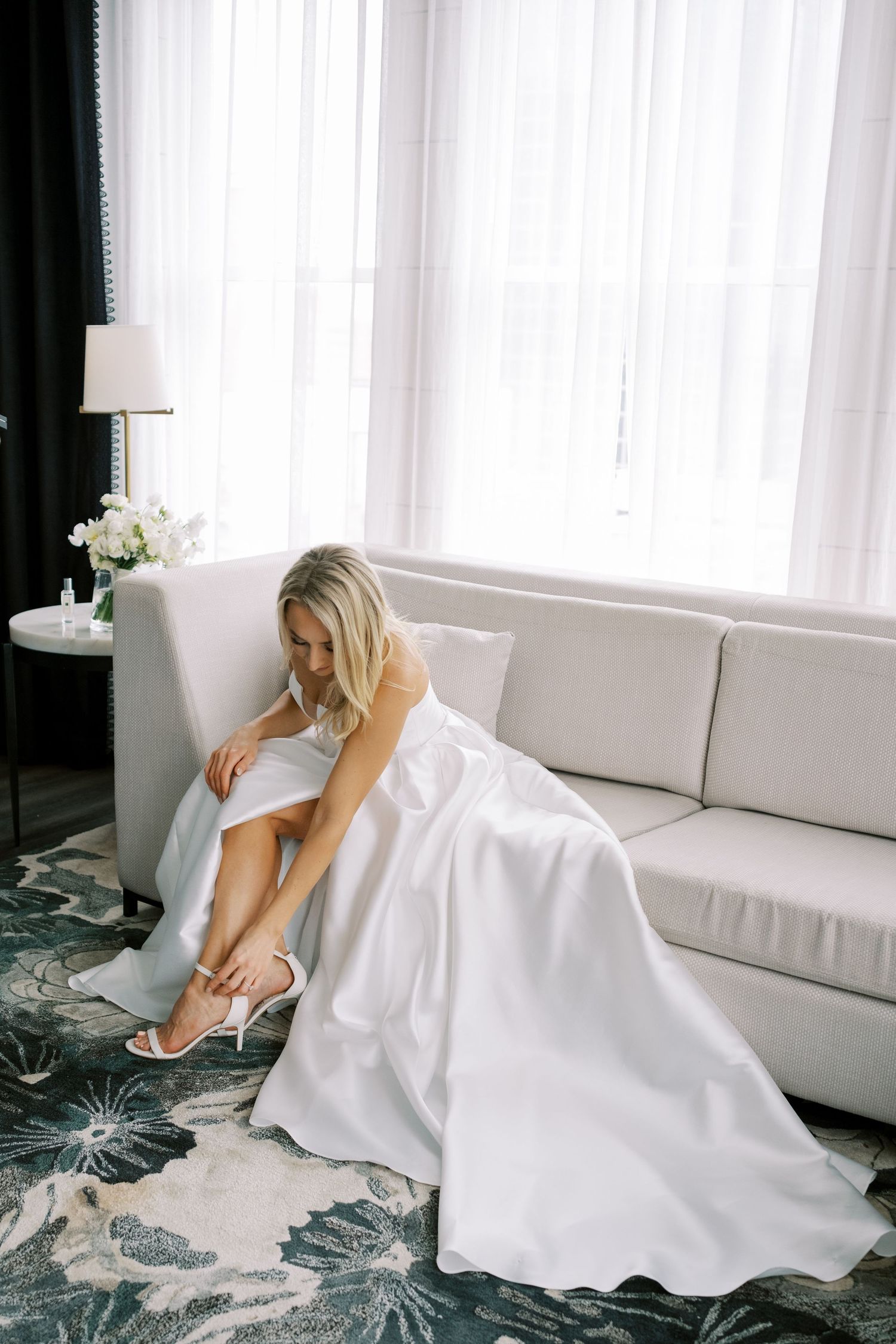 Elegant bride in white gown putting on wedding shoes while seated on gray couch.