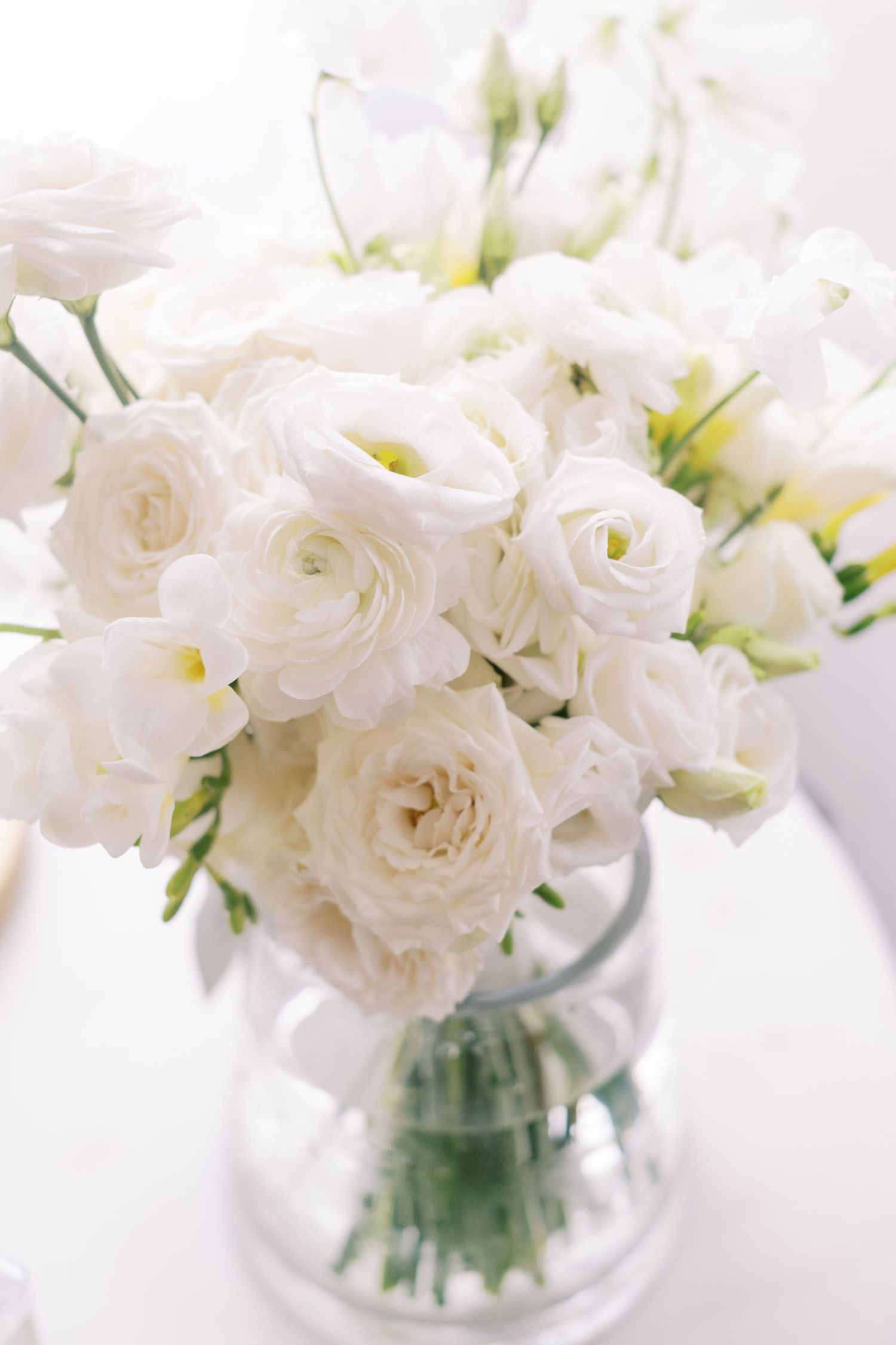 Fresh white roses and ranunculus arranged in clear glass vase against bright background.