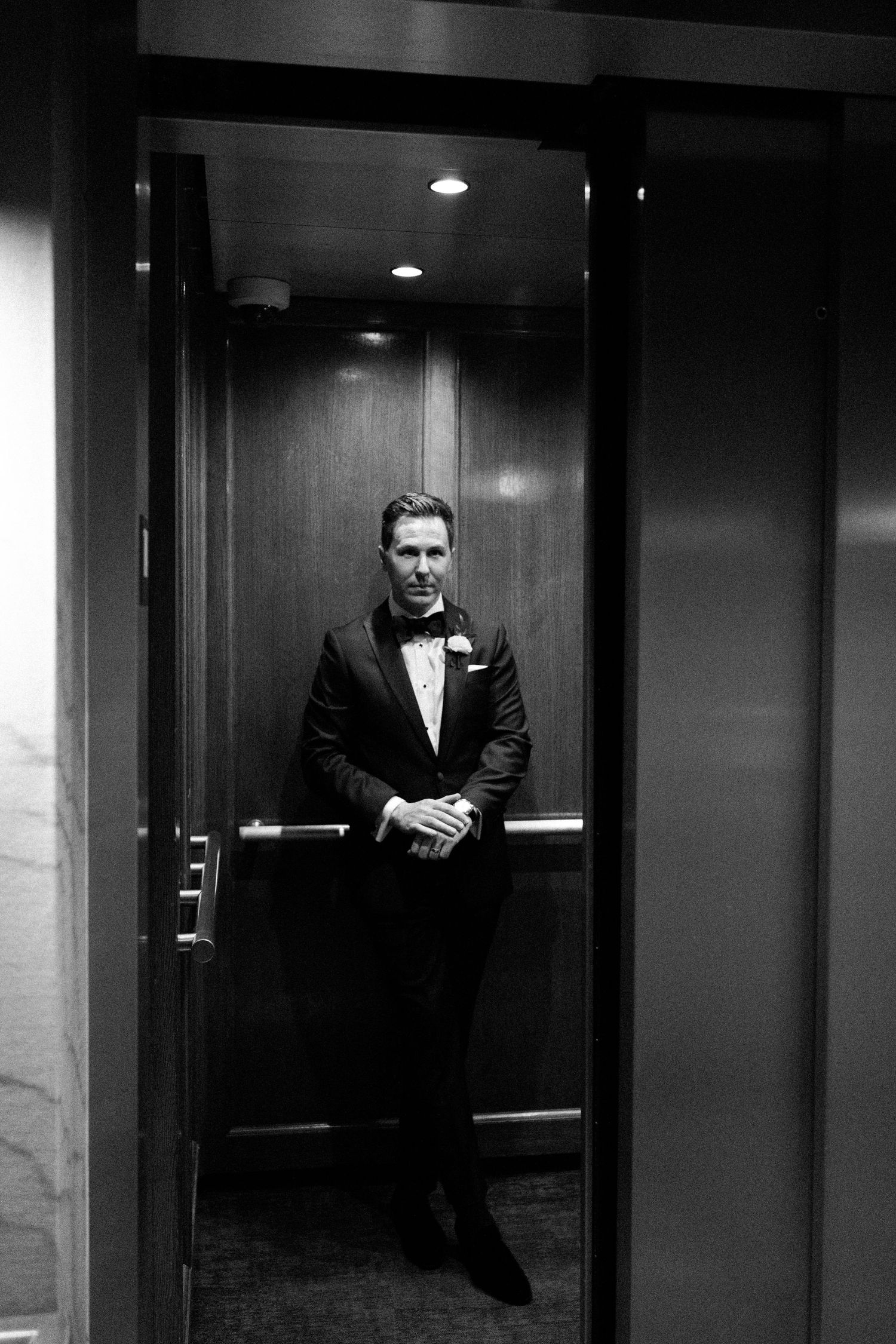 Dramatic black and white portrait of groom in tuxedo standing in modern elevator.
