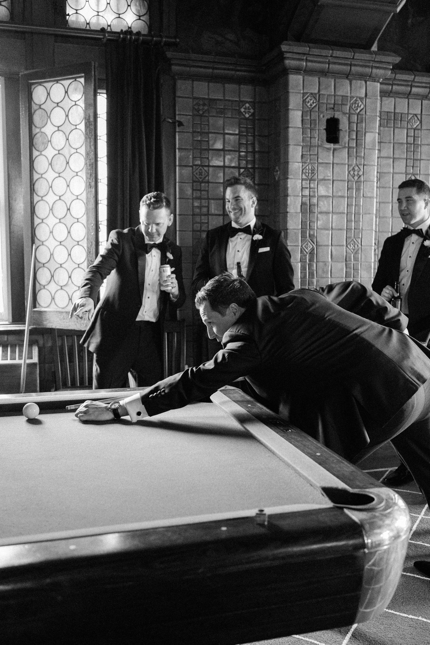 Classic black and white shot of men in tuxedos playing pool in elegant billiards room.