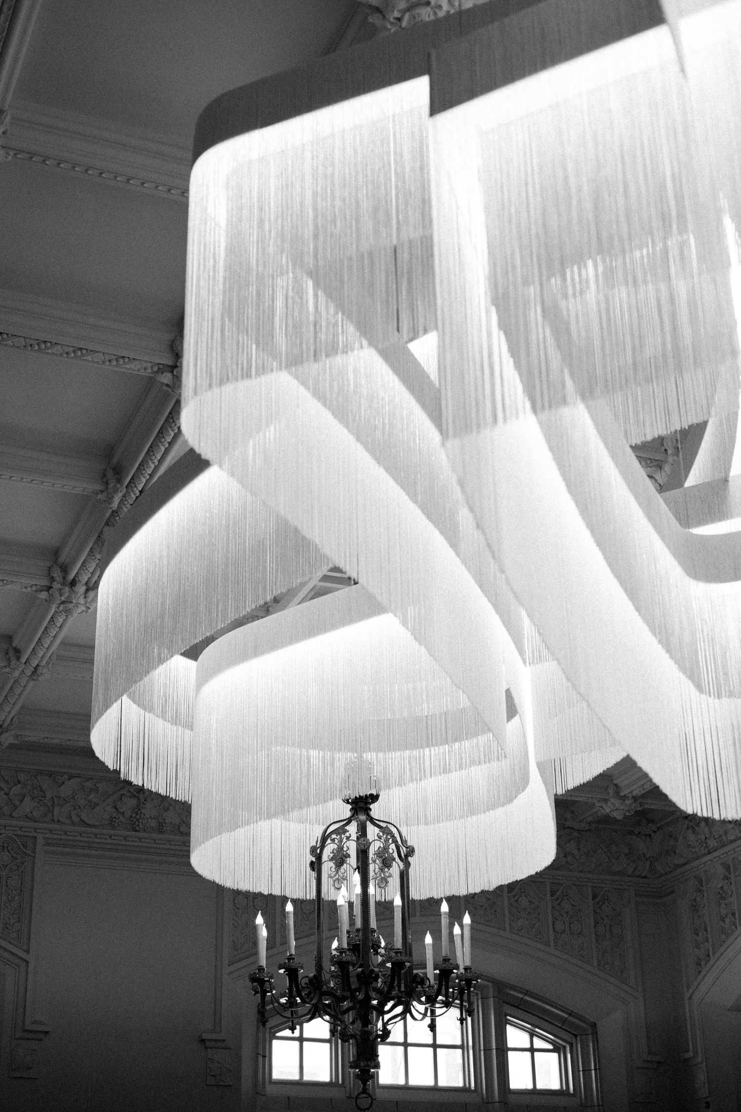 Elegant white fabric chandelier installation with cascading layers hangs above an ornate black candle fixture in a formal room.