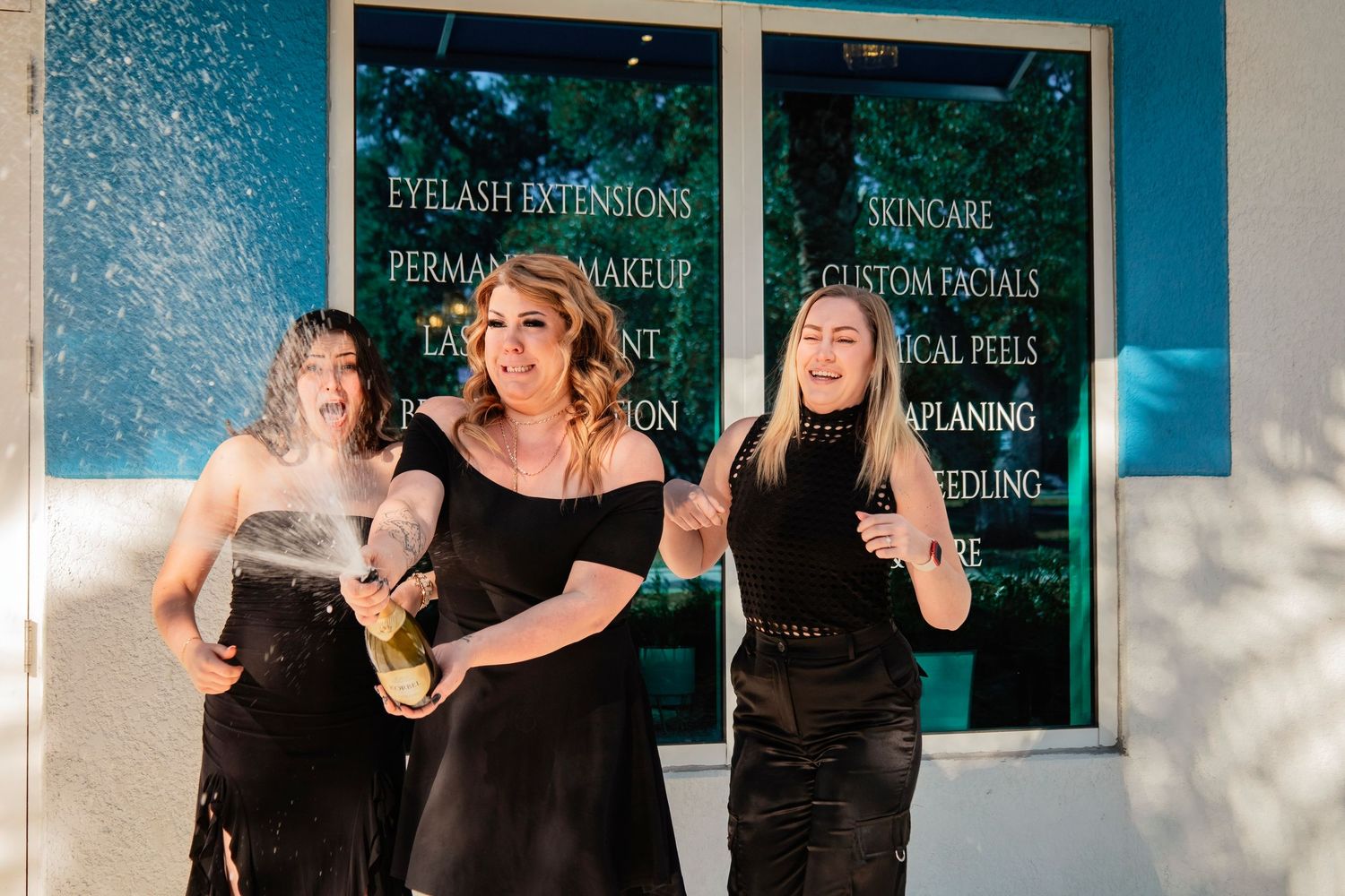 Three women in black outfits toasting and celebrating in front of blue storefront window.