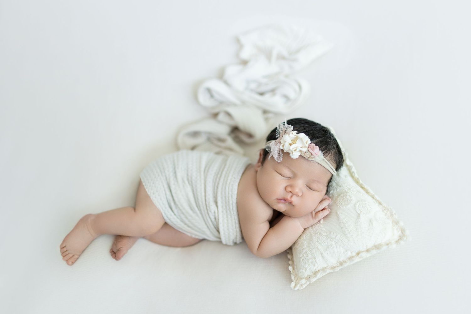 Sleeping newborn wrapped in white fabric on cushion with delicate headpiece. Photos by Tampa Photographer DigitalMyst Photography