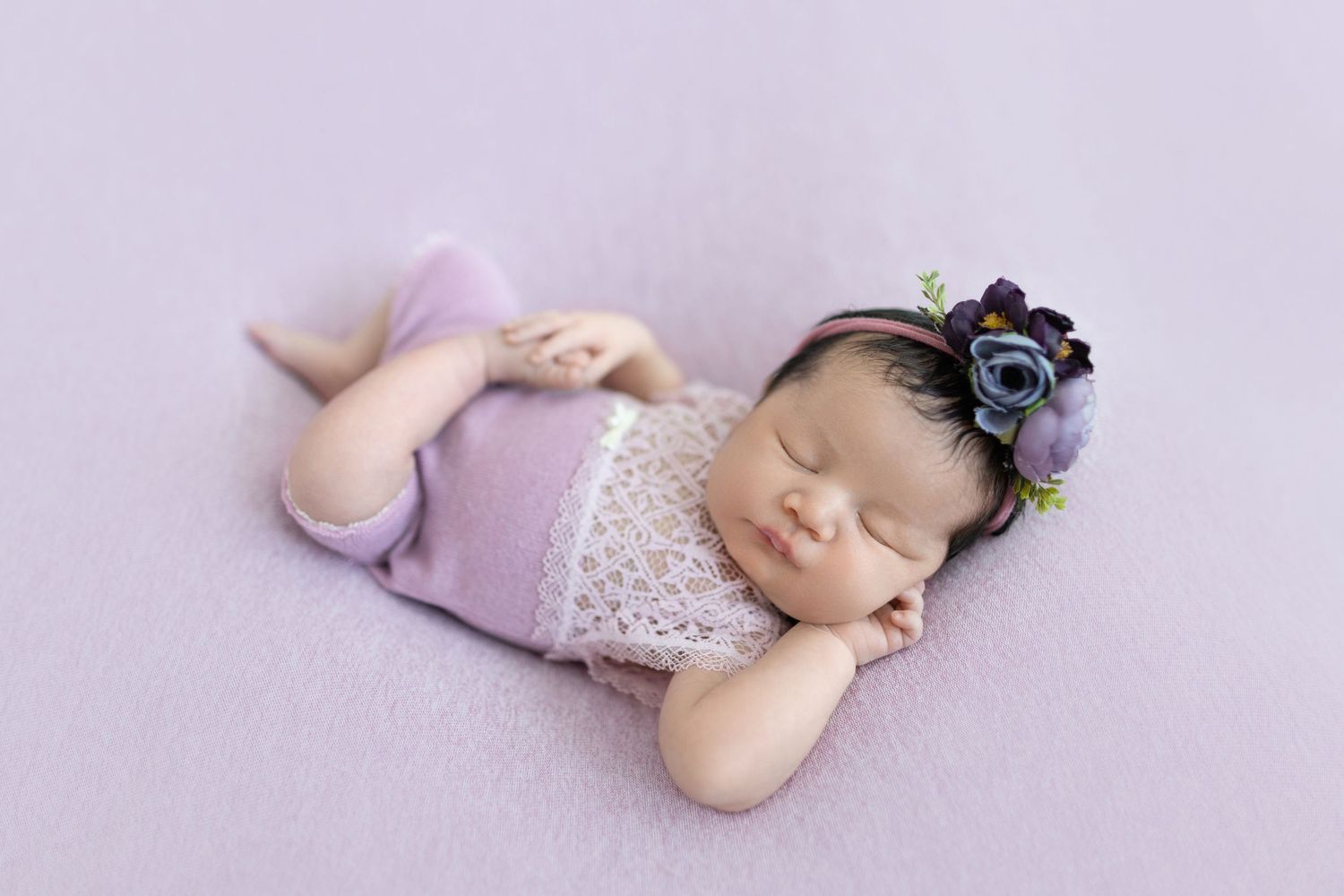 Peaceful sleeping baby in purple outfit with floral headband during newborn photoshoot.