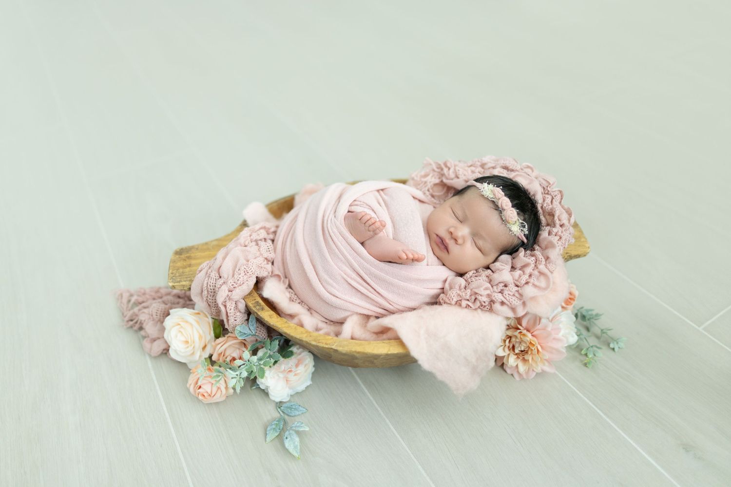 Sleeping newborn wrapped in pink swaddle on wooden bowl with floral decorations.