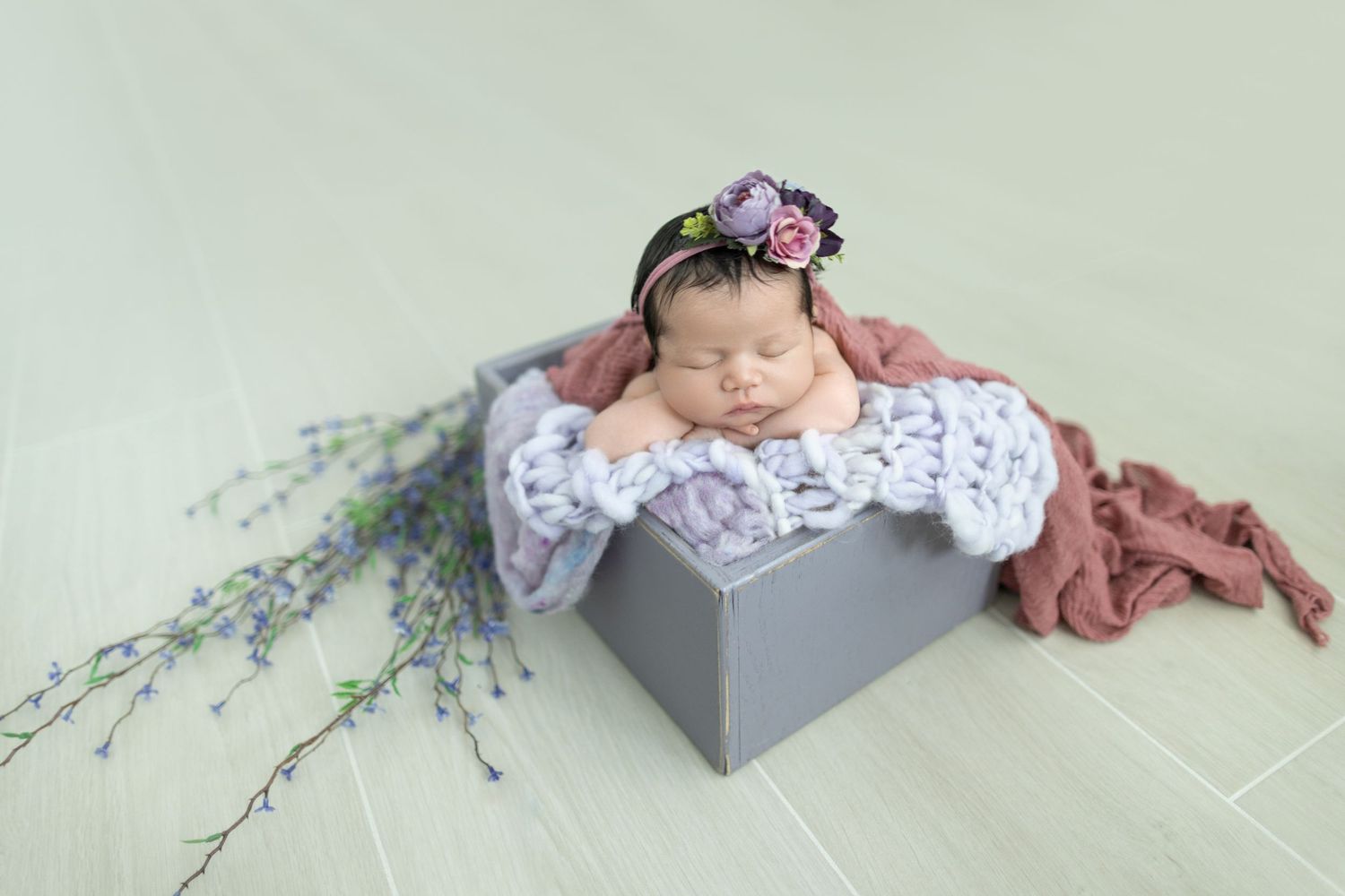 Adorable newborn baby wearing white lace outfit lying on soft grey blanket with delicate floral accents. DigitalMyst Photography