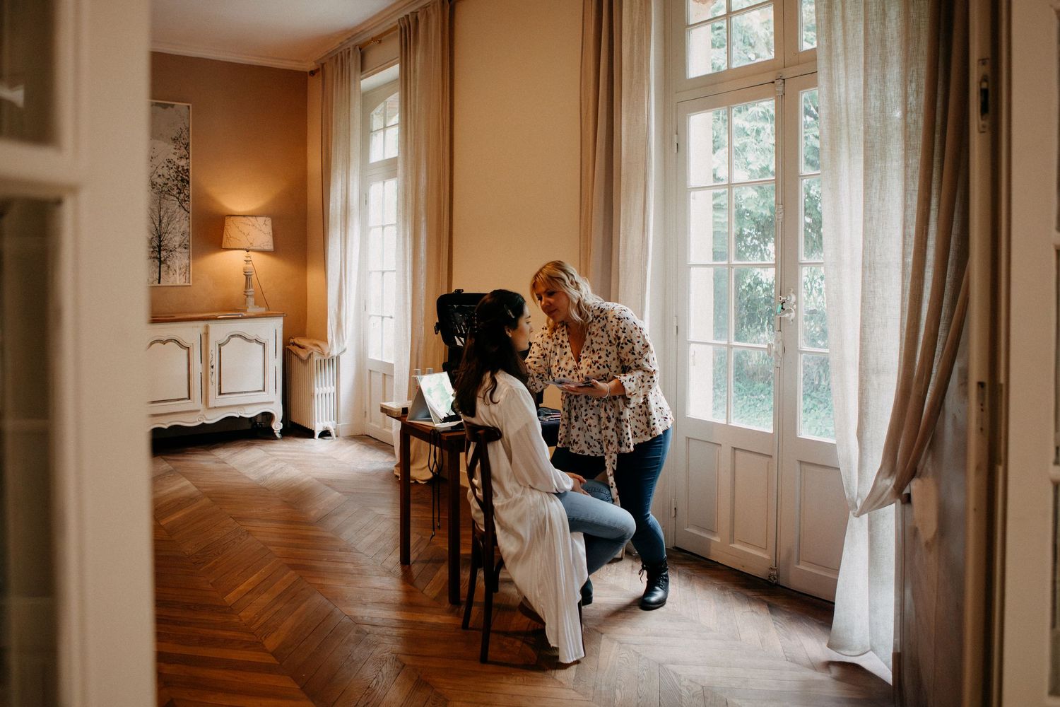 Makeup artist applying makeup to bride seated at vanity in elegant French chateau room.