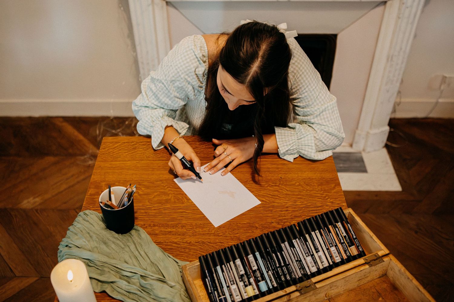 Woman writing calligraphy at a wooden desk with a set of pens and a candle in a cozy interior setting.