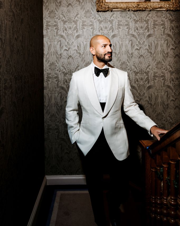 An elegant groom stands in a white tuxedo jacket and bowtie against a patterned wallpaper in a five star hotel.