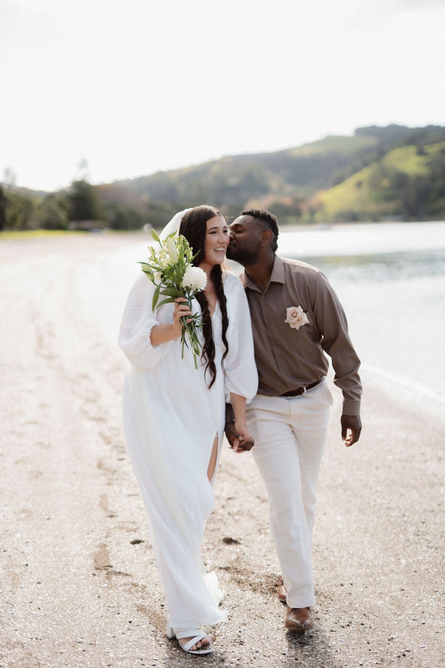 A couple shares an intimate moment while walking along a sandy beach with mountains and water in the background.
