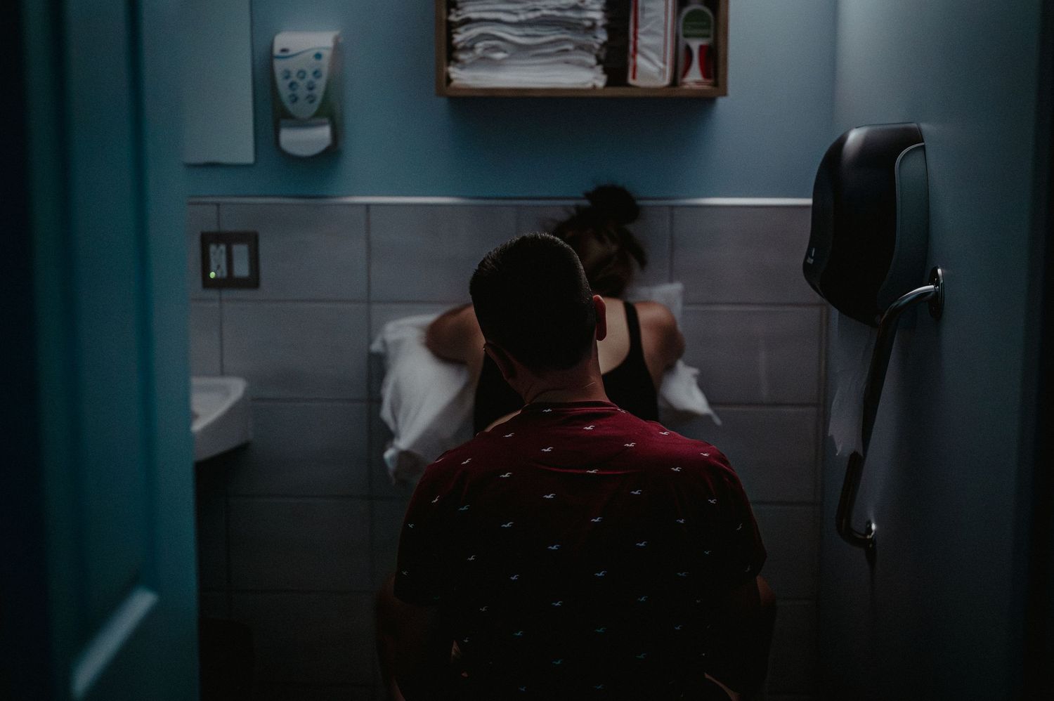 A dark bathroom interior with someone washing their hands at a sink under dim lighting conditions.