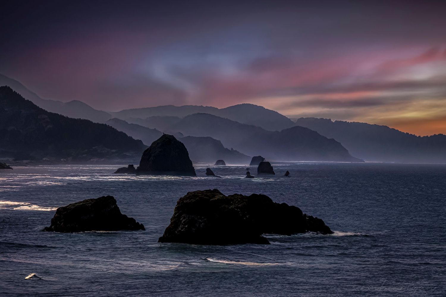 Dramatic sunset over a rugged coastal landscape with rocky cliffs and crashing waves along a misty shoreline.