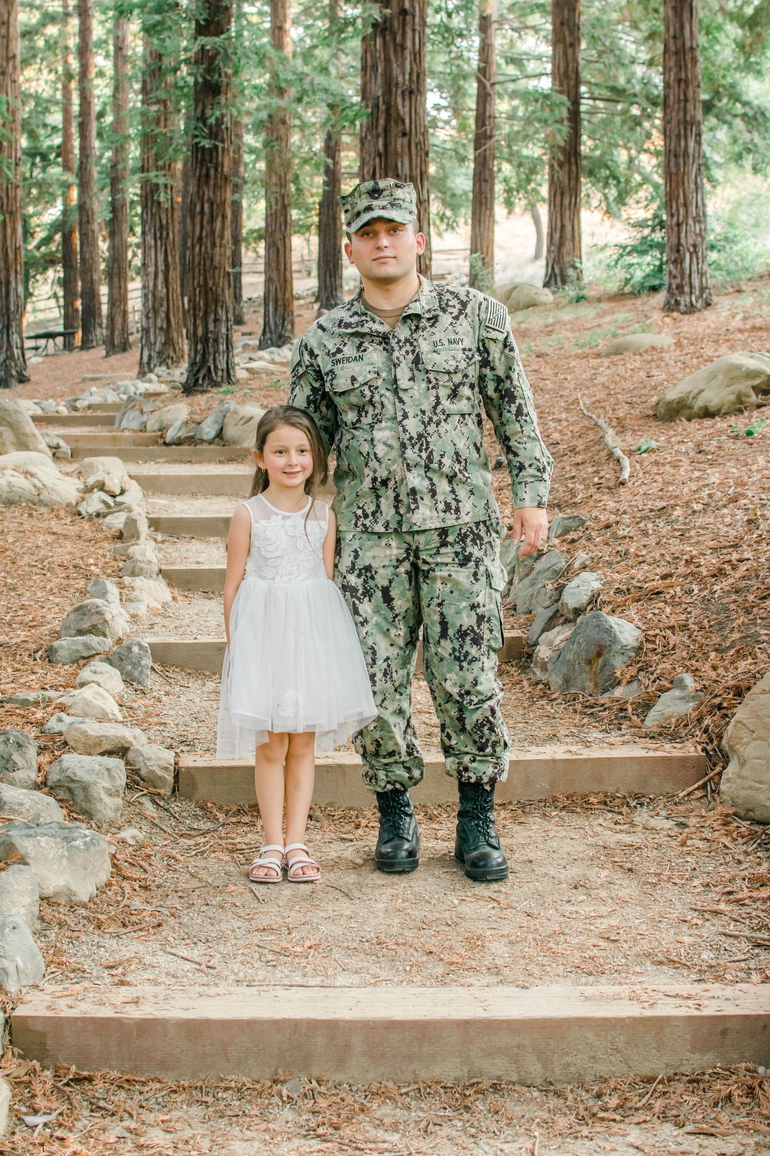 Military personnel and child standing together on stone steps in a wooded forest setting.