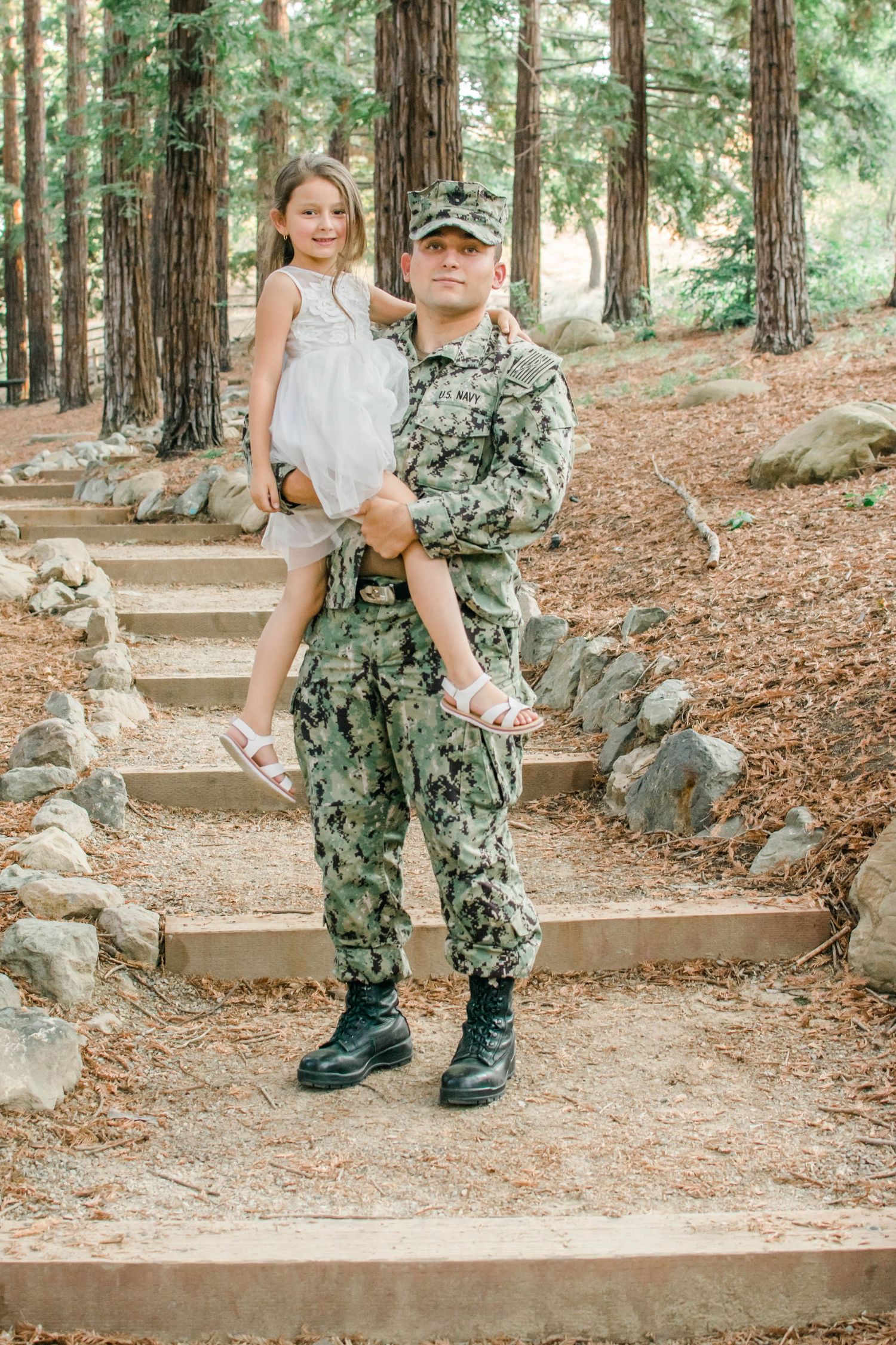 A service member in camouflage uniform stands with a small companion on outdoor stone steps.