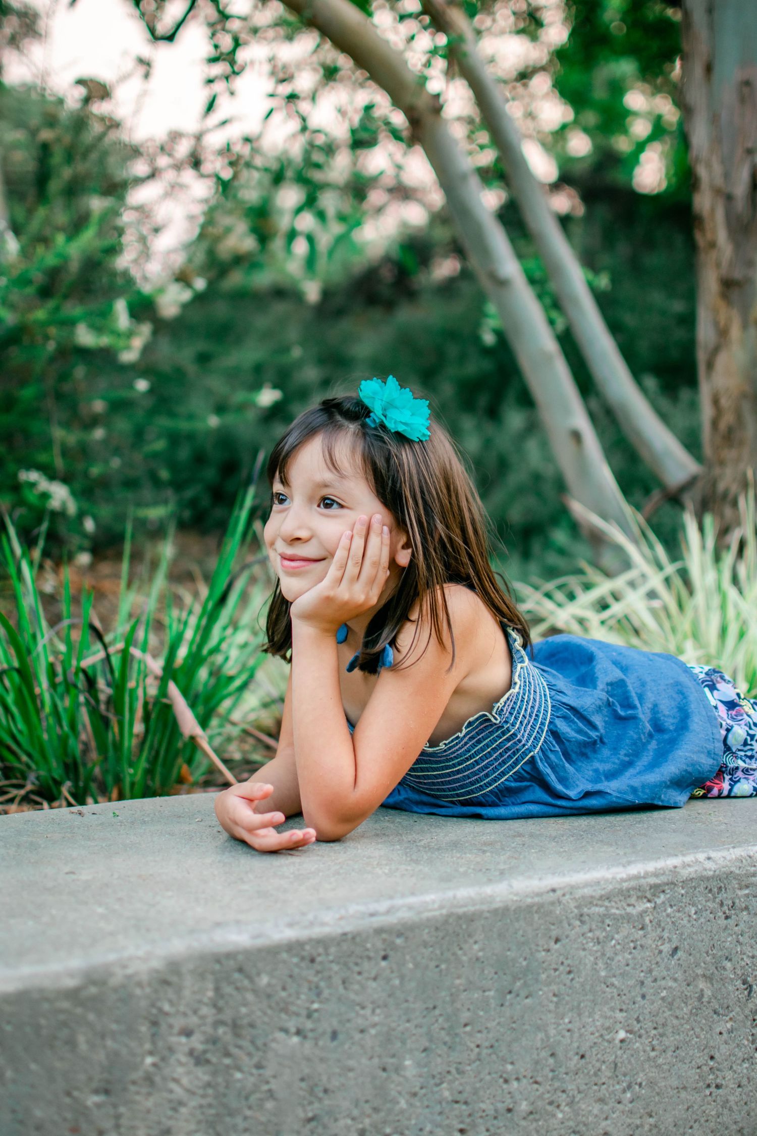 Series of relaxed poses on a concrete surface with blue bow and denim outfit against green foliage.