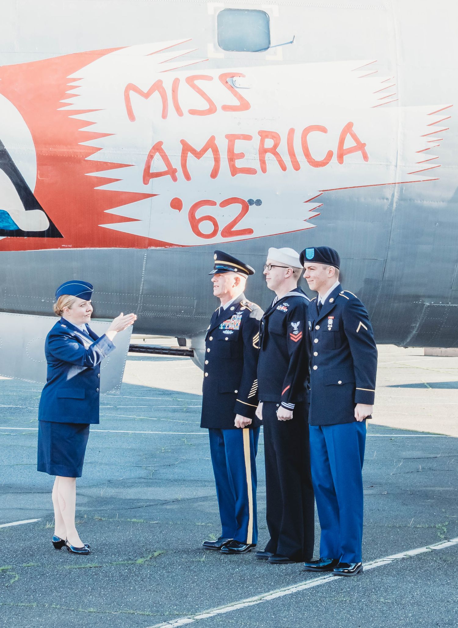 Military personnel in dress uniforms saluting near an aircraft with 'Miss America' text visible.