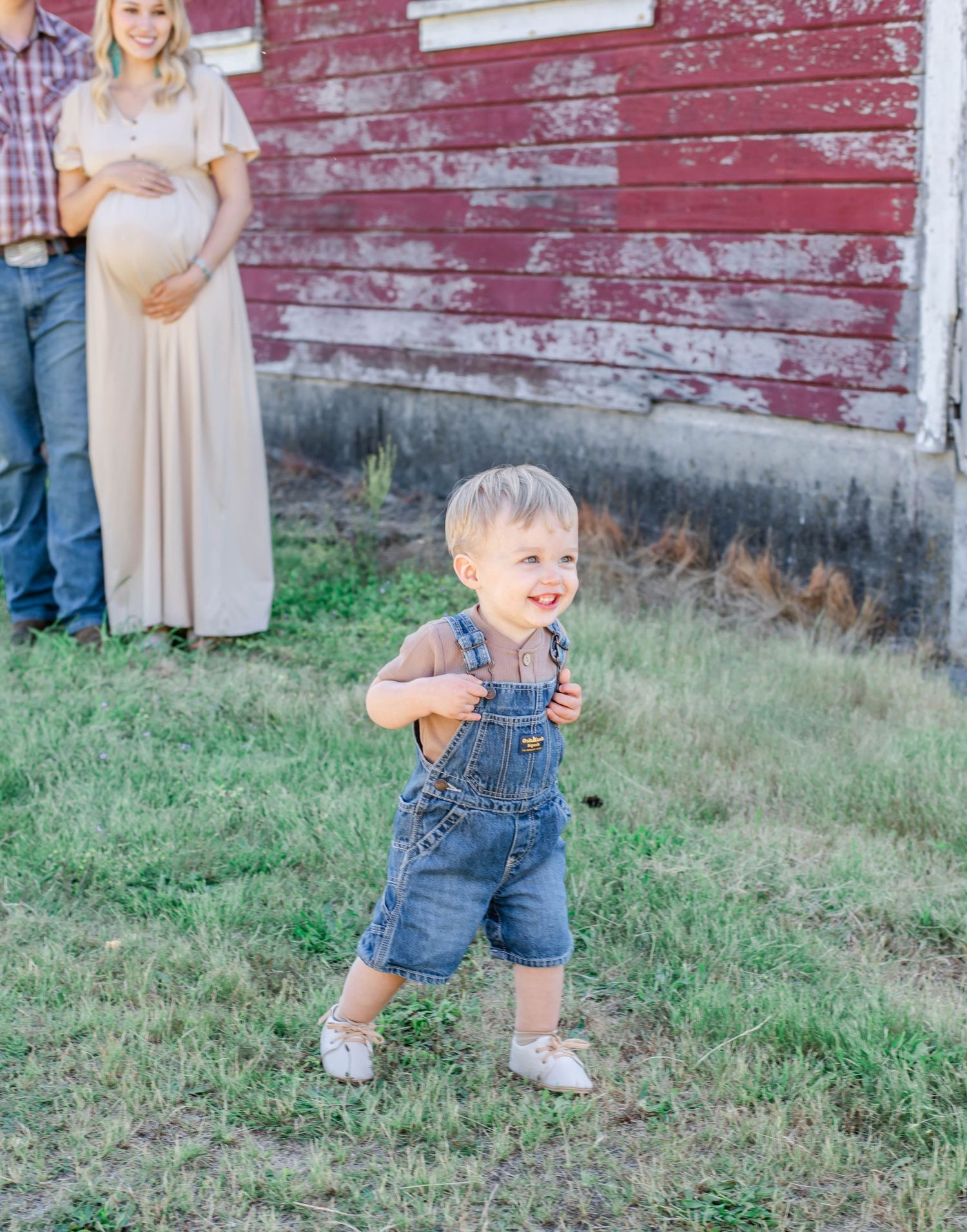 A toddler in denim overalls plays in the grass near a rustic red barn.
