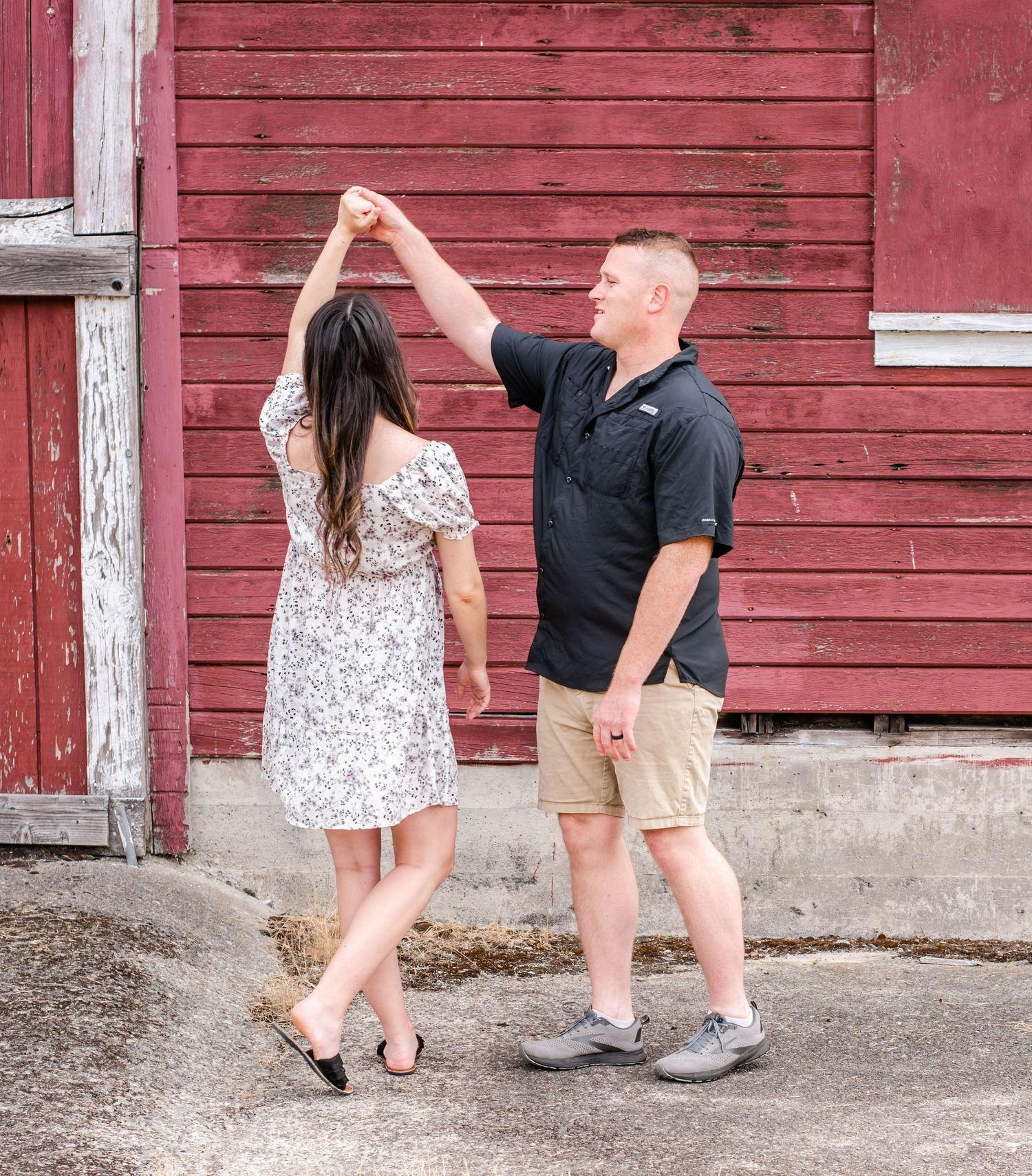 A couple dances playfully together beside a weathered red barn during their photoshoot.