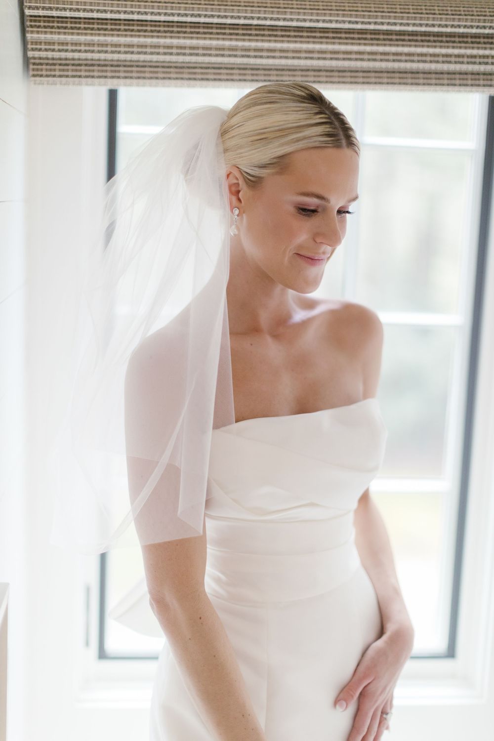 A bride in an elegant strapless white wedding dress and veil looks down with a gentle smile near a window.