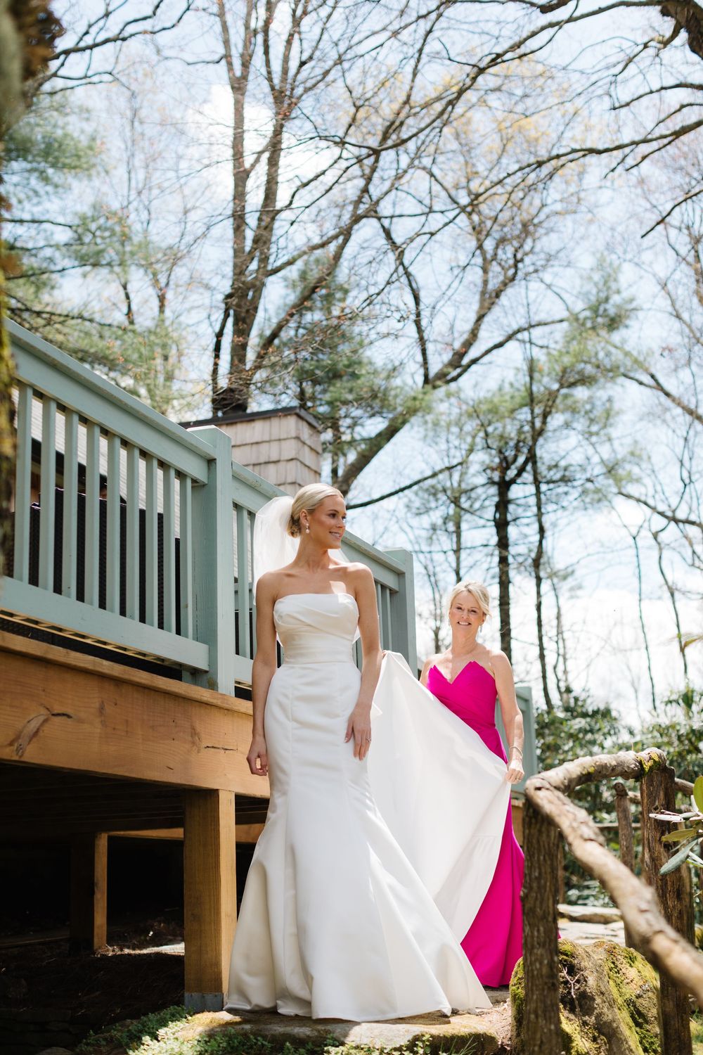 Two people in formal attire stand together on a wooden deck surrounded by tall trees on a sunny day.