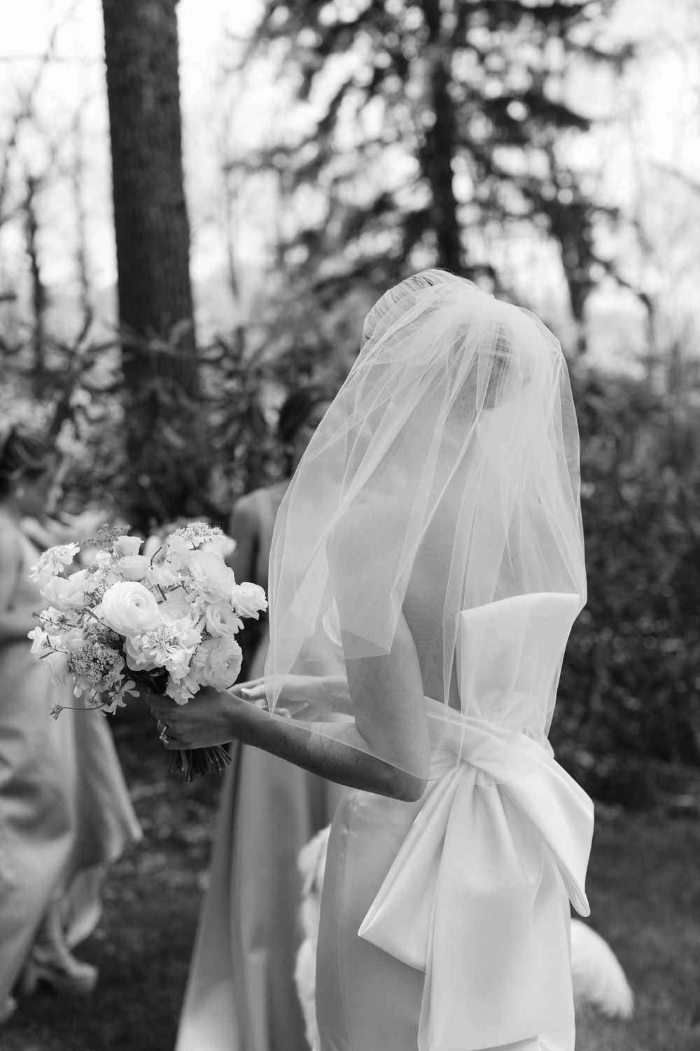 A bride in an elegant white dress with a large bow and veil holds a floral bouquet in a forest setting, captured in black and white.