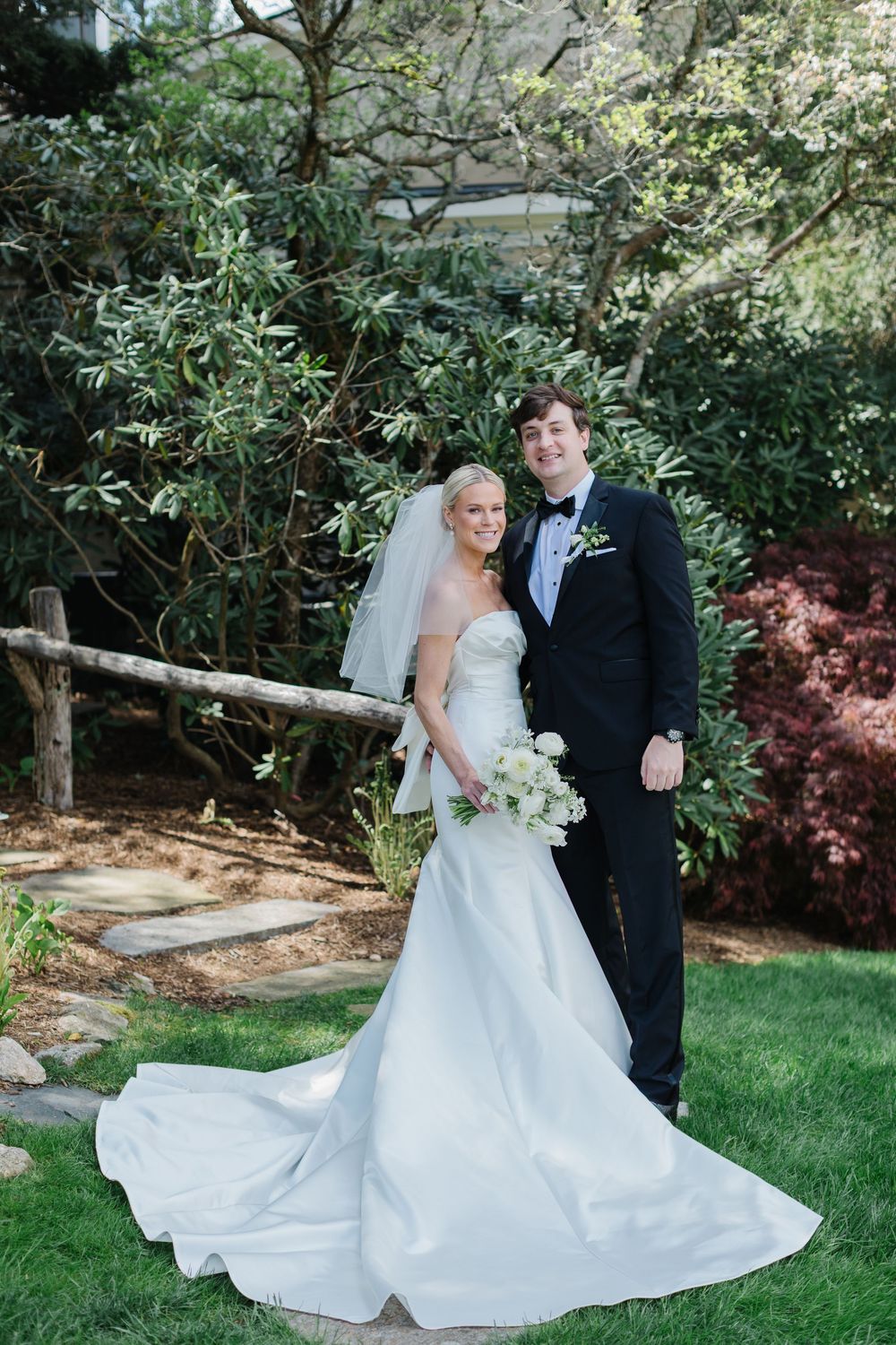 A couple poses for a formal wedding photo in a garden setting with lush greenery and a rustic wooden fence.