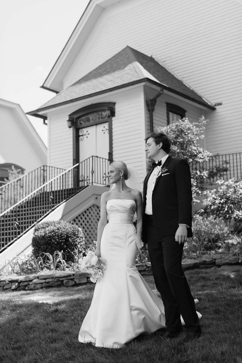 Newlywed couple in formal wedding attire standing on lawn in front of white church entrance with stairs.