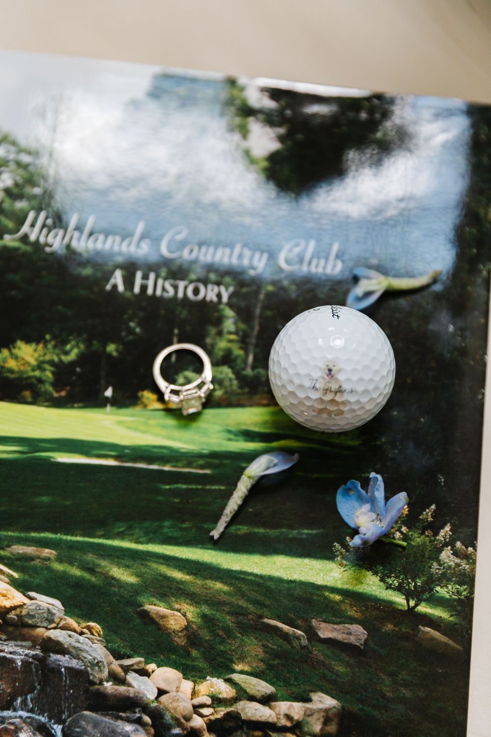 A golf ball and tee rest on a book titled 'Highlands Country Club: A History' with a blue flower in the corner.