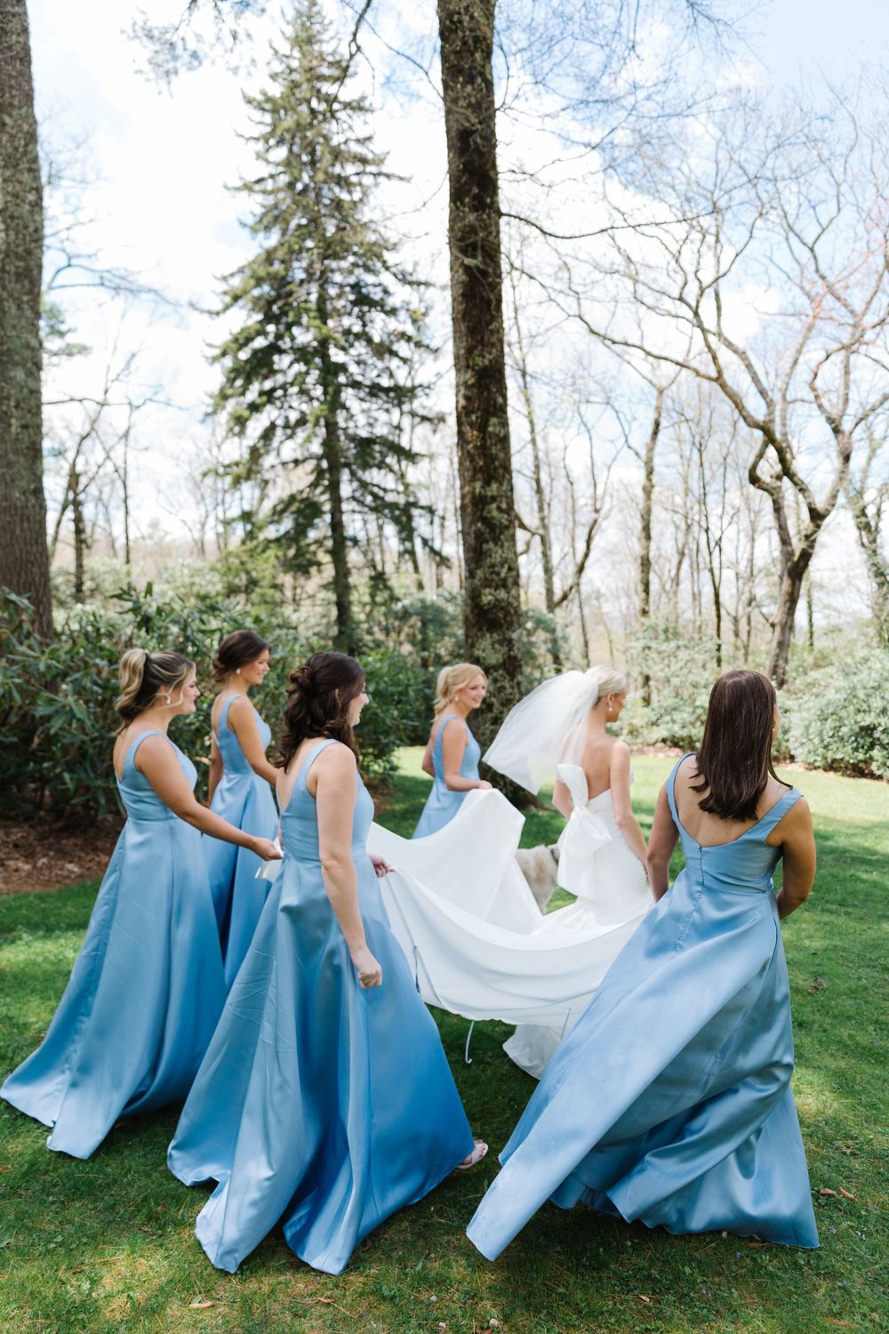 Bridesmaids in matching light blue dresses help fluff a white wedding gown in an outdoor spring garden setting.