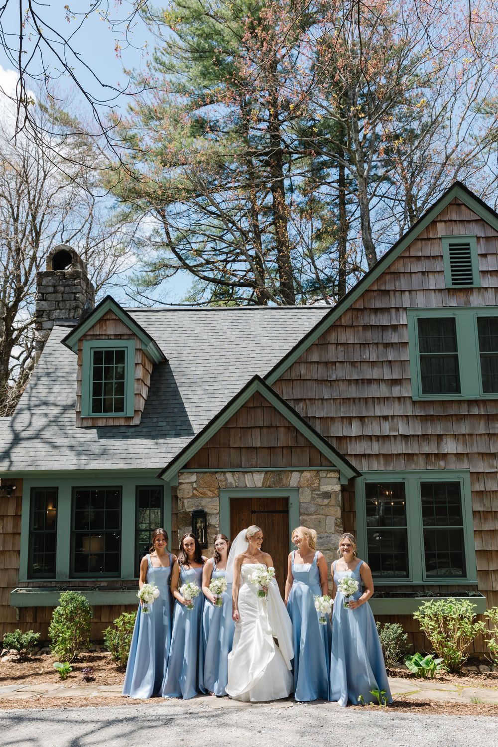 Wedding party stands in front of rustic cedar shake home with stone facade and green trim on a sunny day.