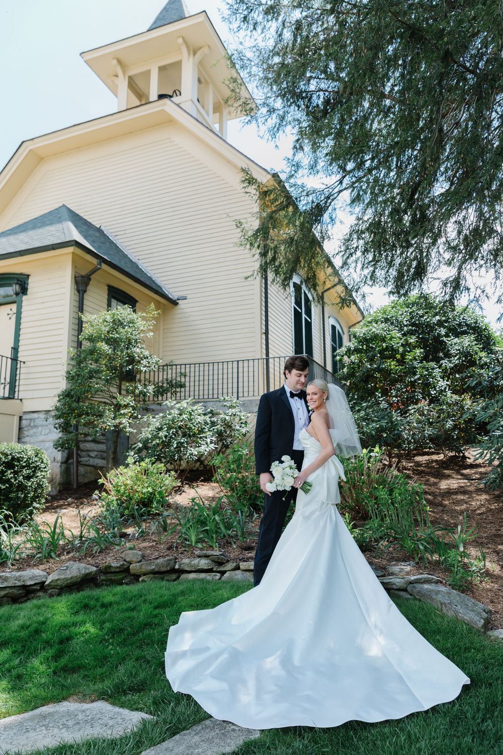 A couple in wedding attire stands close together outside a white church building with a lush garden landscape. highlands north carolina