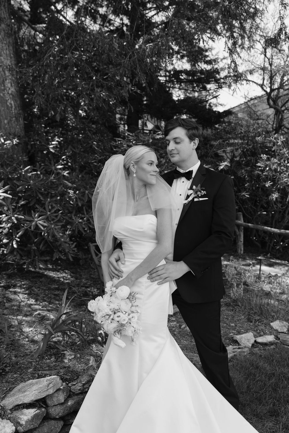 A black and white wedding photo of a couple embracing in a garden setting with the bride in a white gown holding flowers.